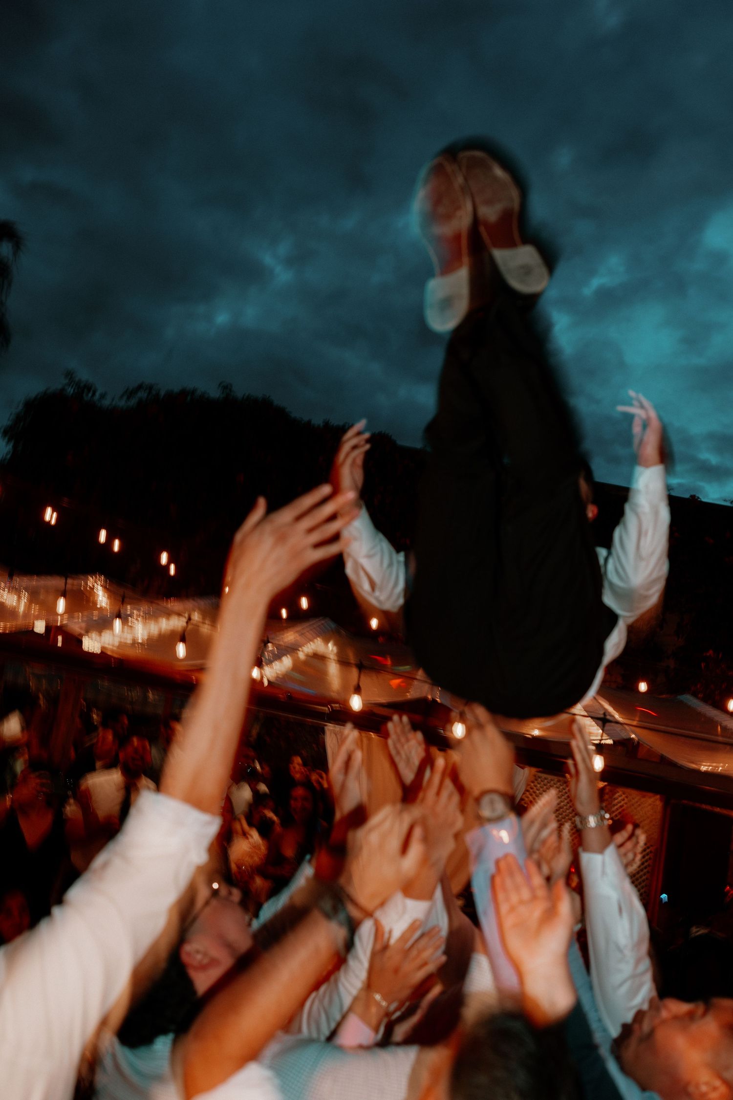 Crowd lifting a person in the air at an outdoor evening wedding celebration under dramatic cloudy skies.