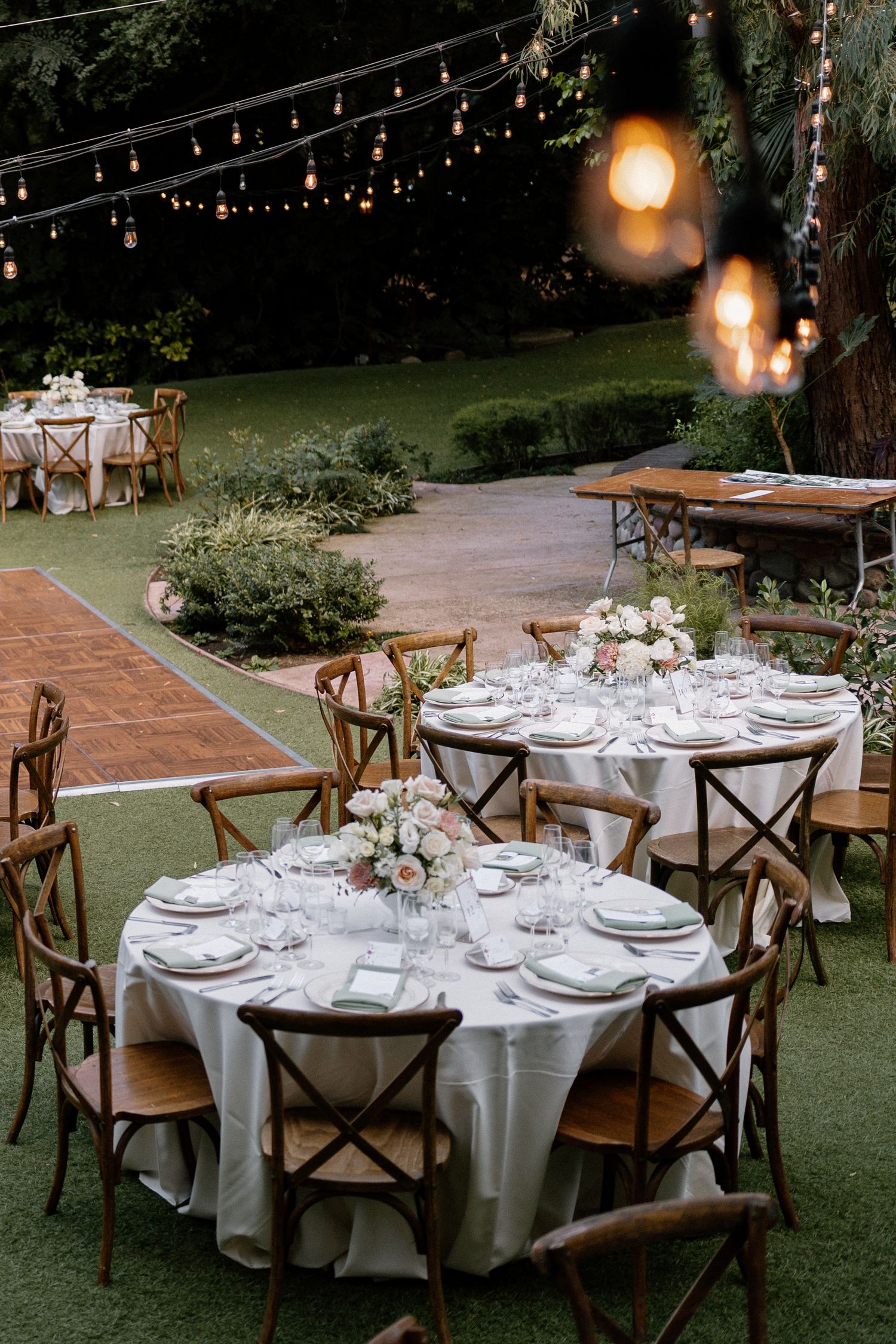 Elegant outdoor wedding reception with round tables, cross-back chairs, floral centerpieces, and string lights.