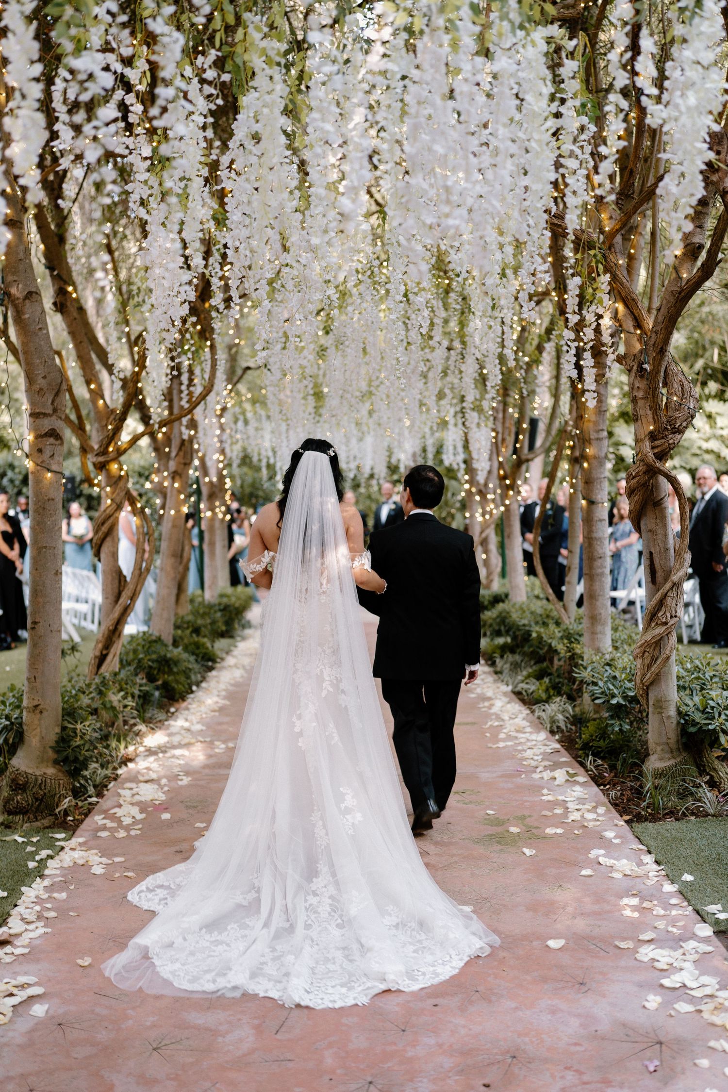 Bride in lace gown with cathedral veil walks with groom under white wisteria-draped trees at outdoor wedding.