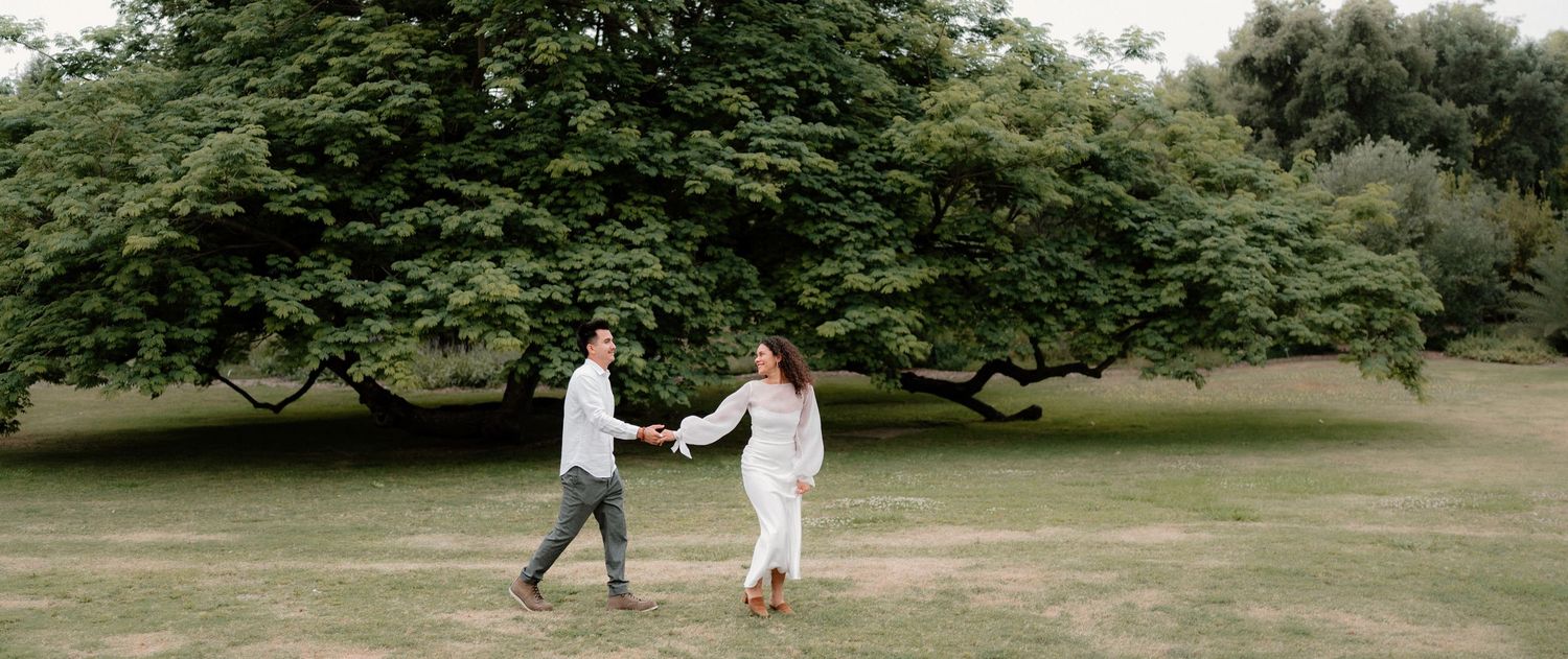 Couple holding hands and walking through a lush green park during an engagement photo session.
