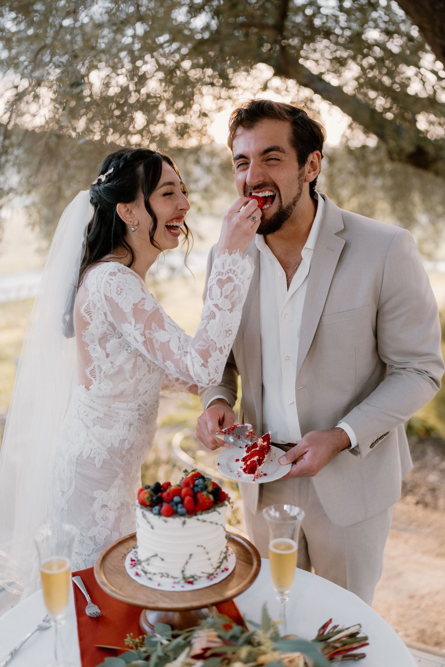Laughing bride feeding groom a piece of red velvet wedding cake at outdoor garden reception.