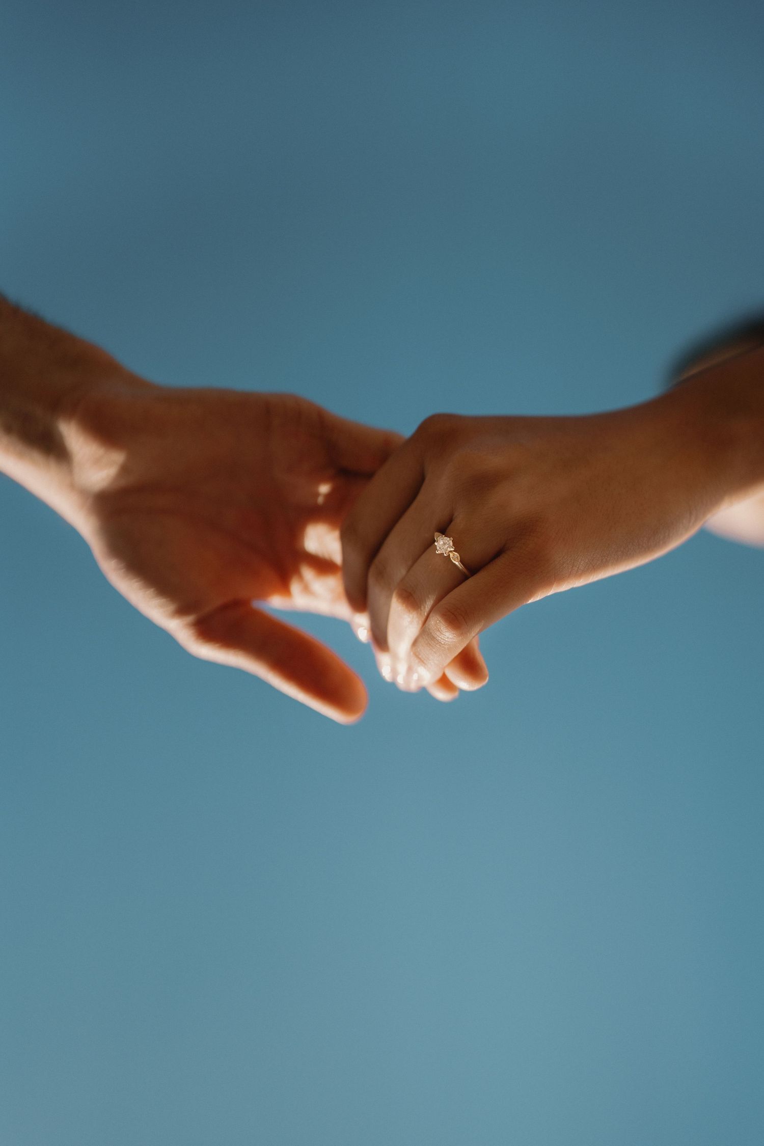 Close-up of couple's hands touching with a delicate diamond engagement ring against a blue sky.