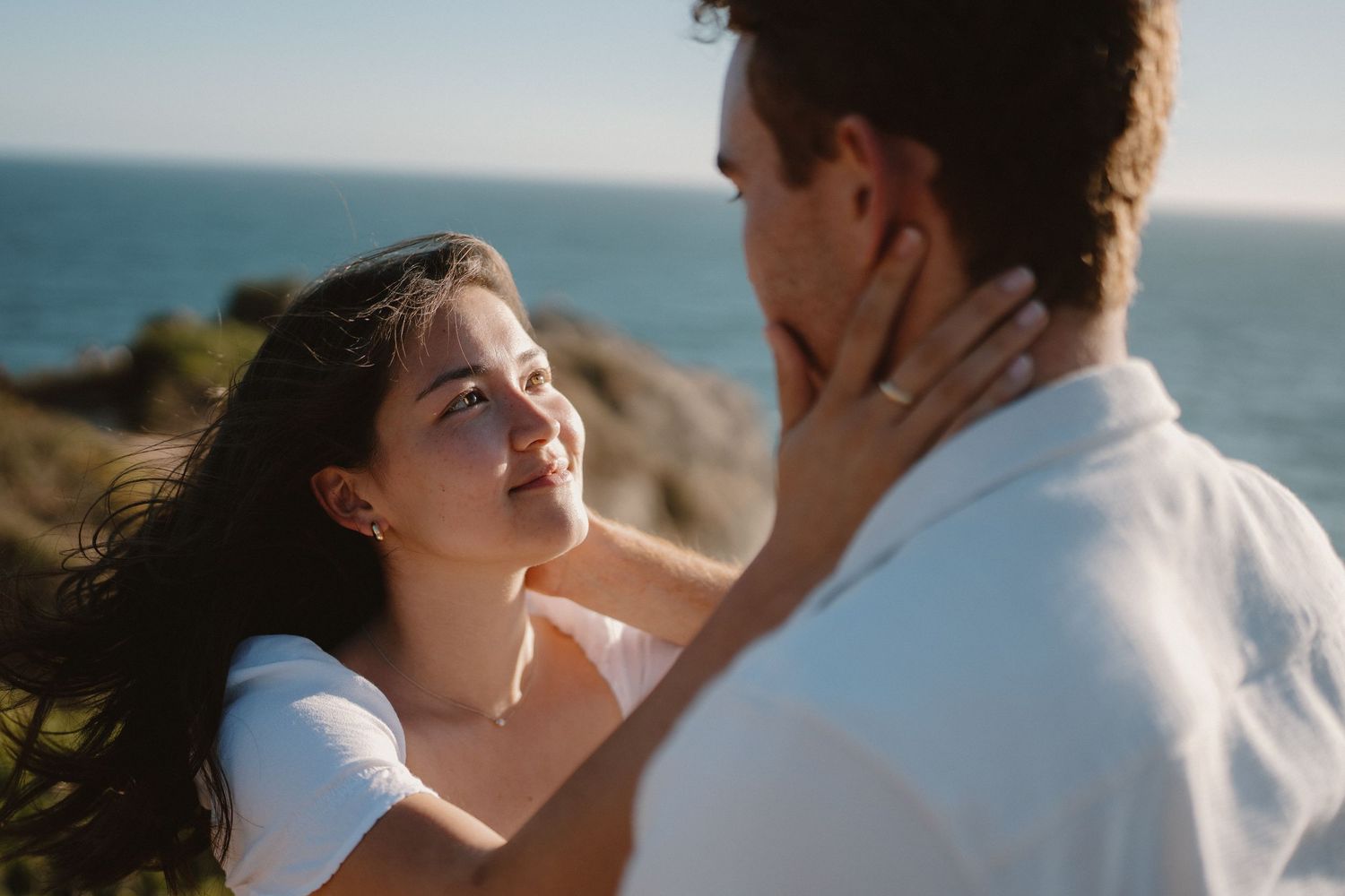 Romantic couple sharing a tender moment by the ocean, woman gently touching man's face in golden sunlight.
