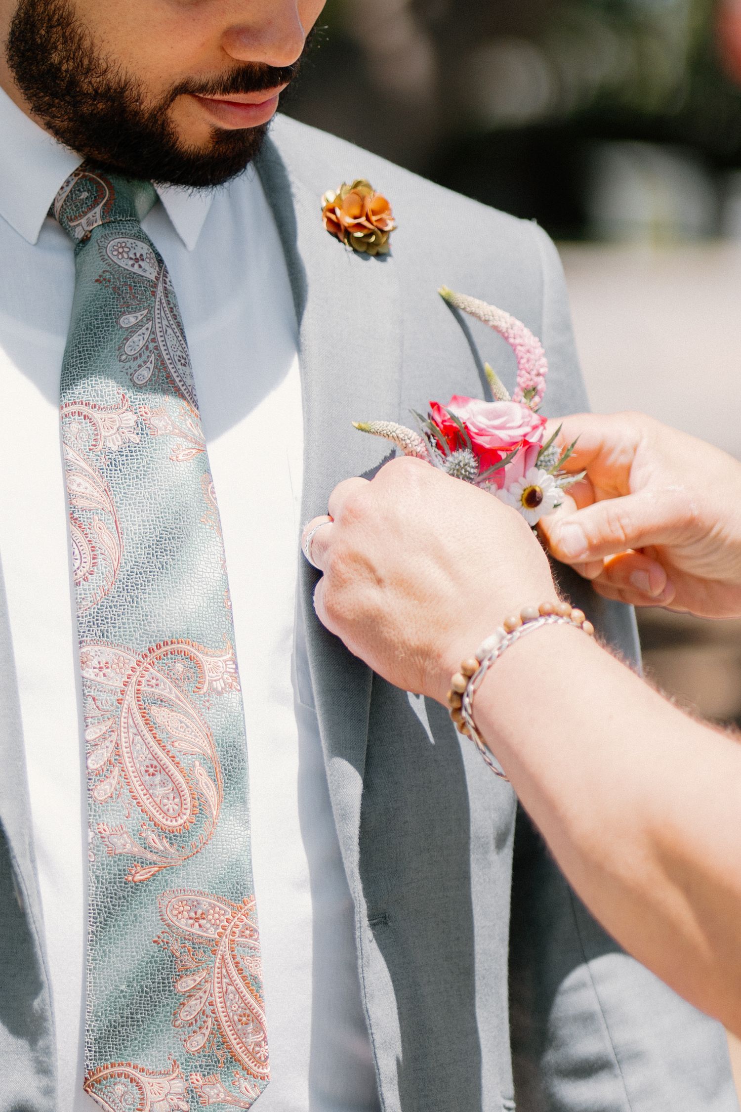 Groom in gray suit with paisley tie getting boutonniere pinned on lapel.