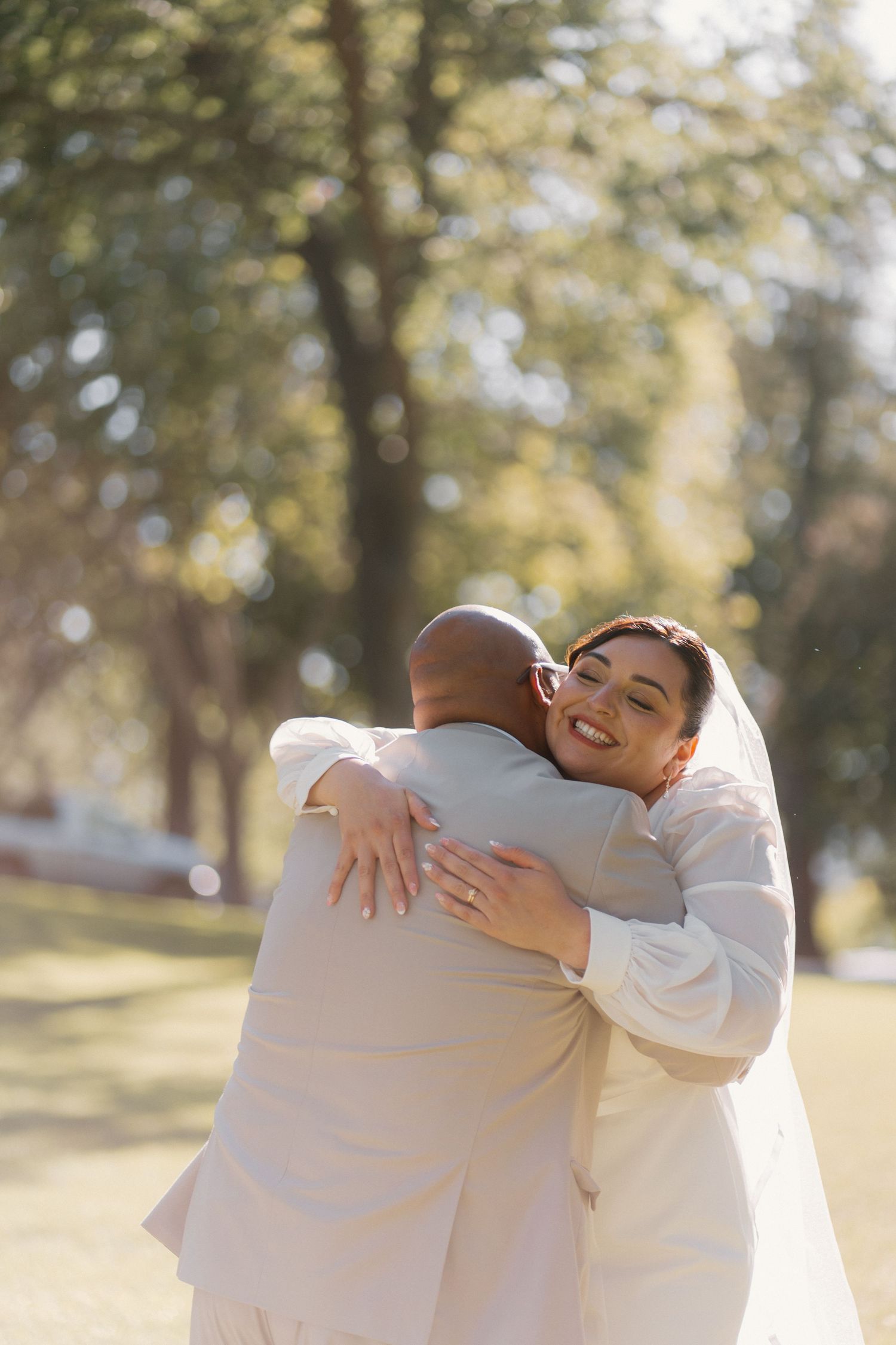Smiling bride in white wedding dress and veil embracing groom outdoors in a sunny park setting.