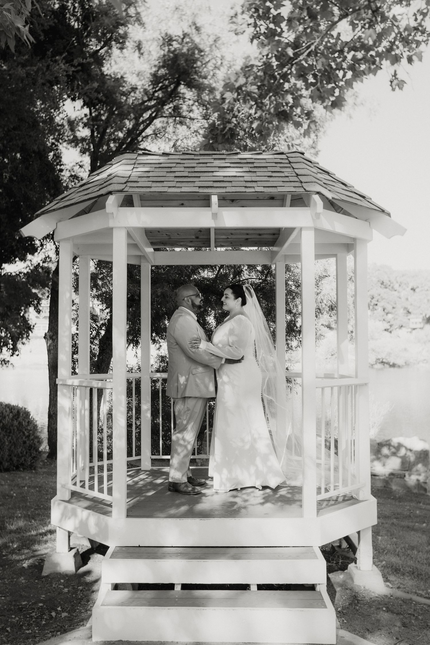 Bride and groom share romantic moment inside white gazebo surrounded by trees in black and white.