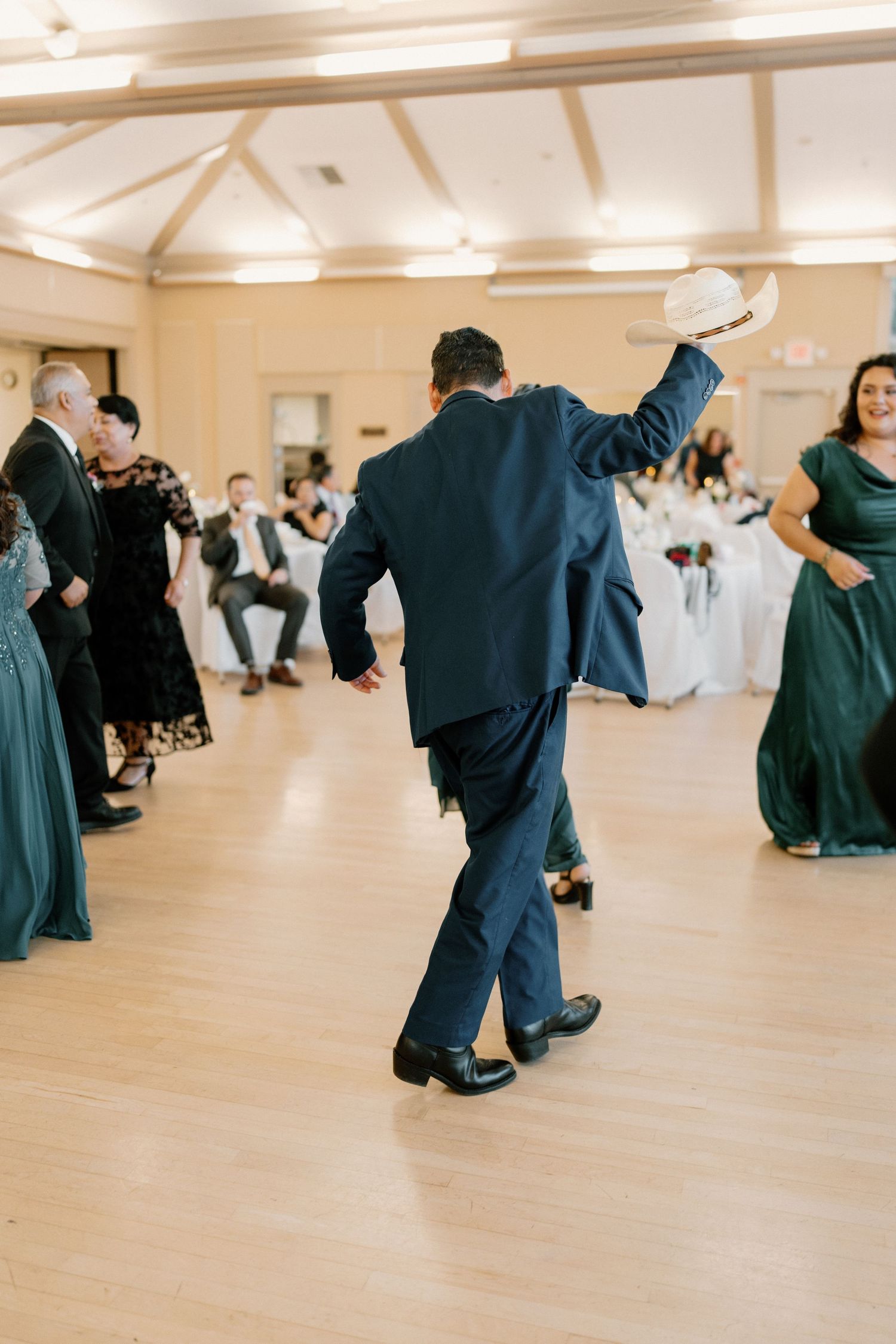 Guest in navy suit dancing with white hat raised at lively wedding reception on dance floor.