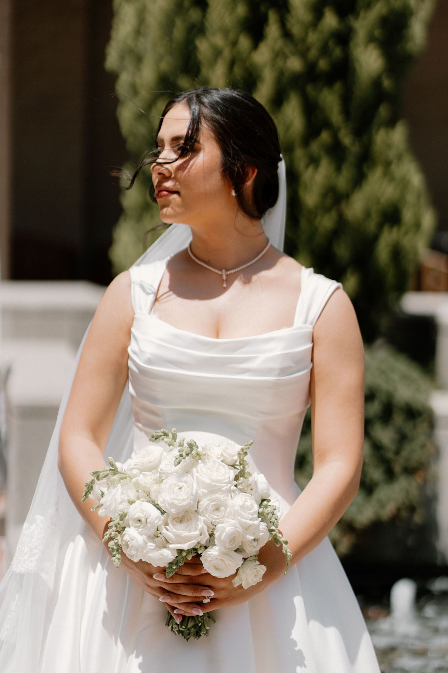 Bride in white satin gown with veil and glasses holds white rose bouquet outdoors in sunlight.