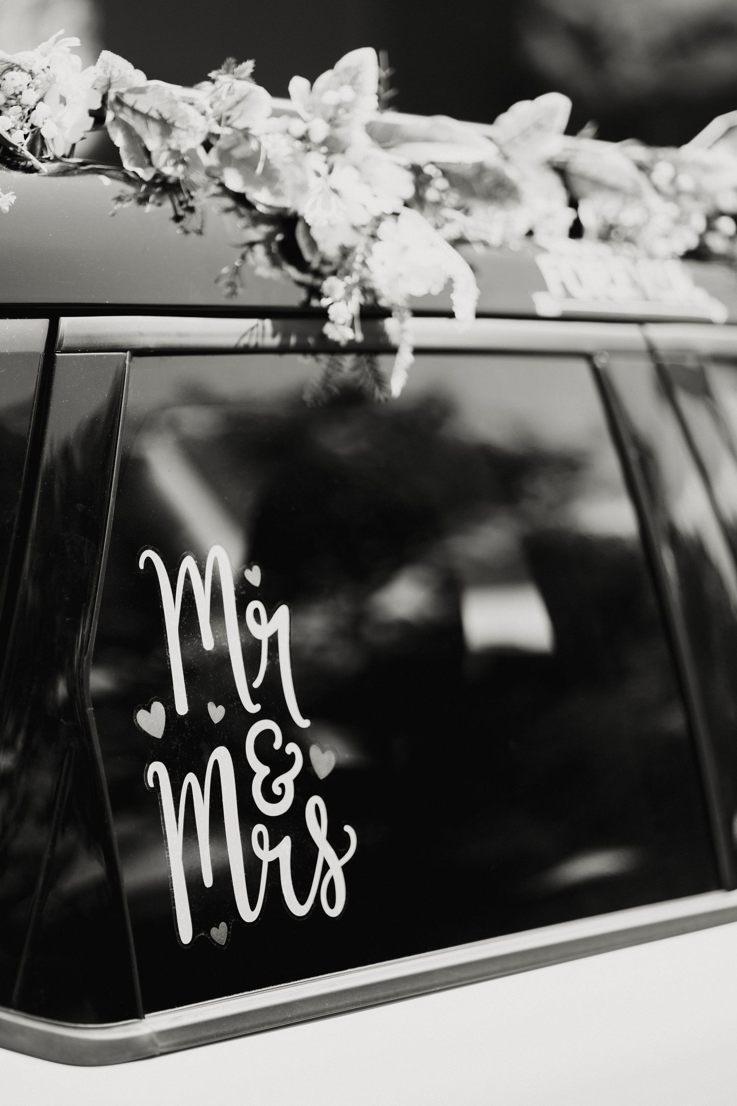 Black and white photo of a wedding car window with Mr and Mrs decal and floral roof decorations.