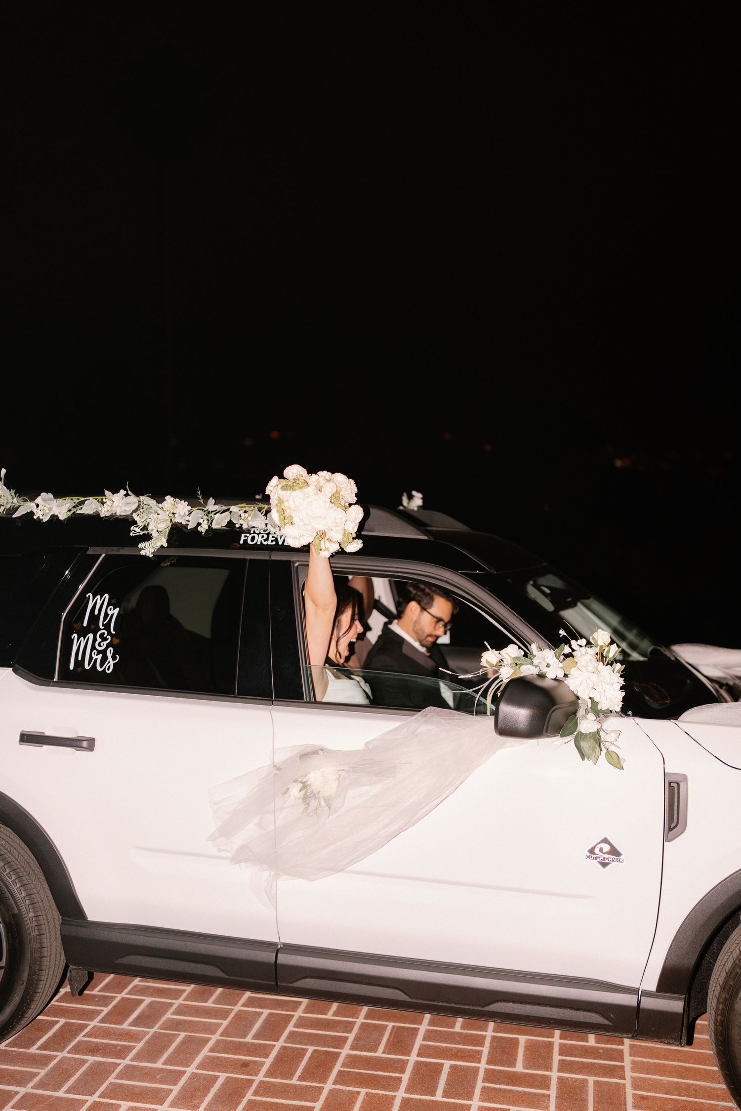 Newlyweds departing in a white SUV decorated with Mr & Mrs decal and white floral arrangements.