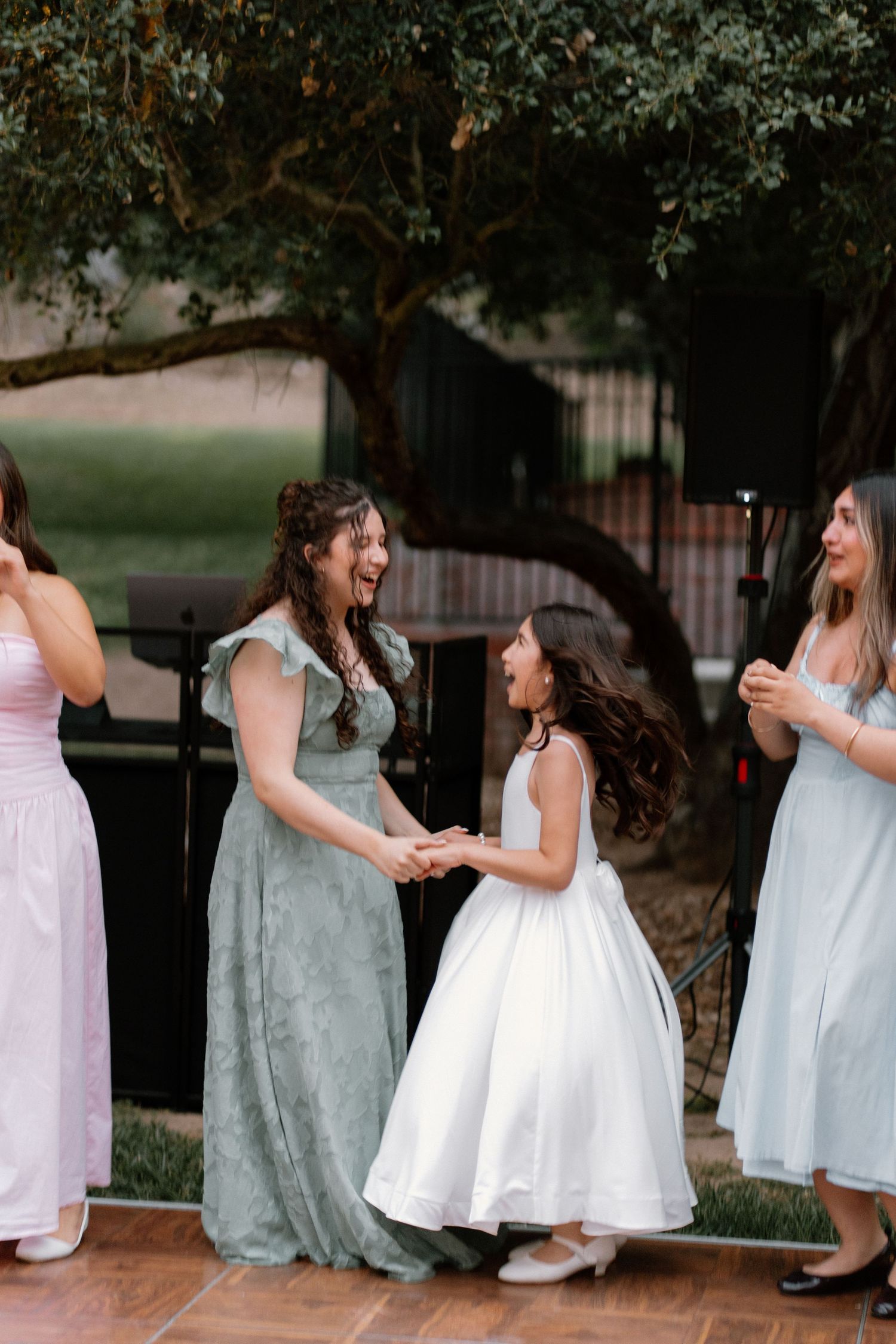 Bridesmaids in sage and pink dresses dance joyfully with flower girl at outdoor wedding.