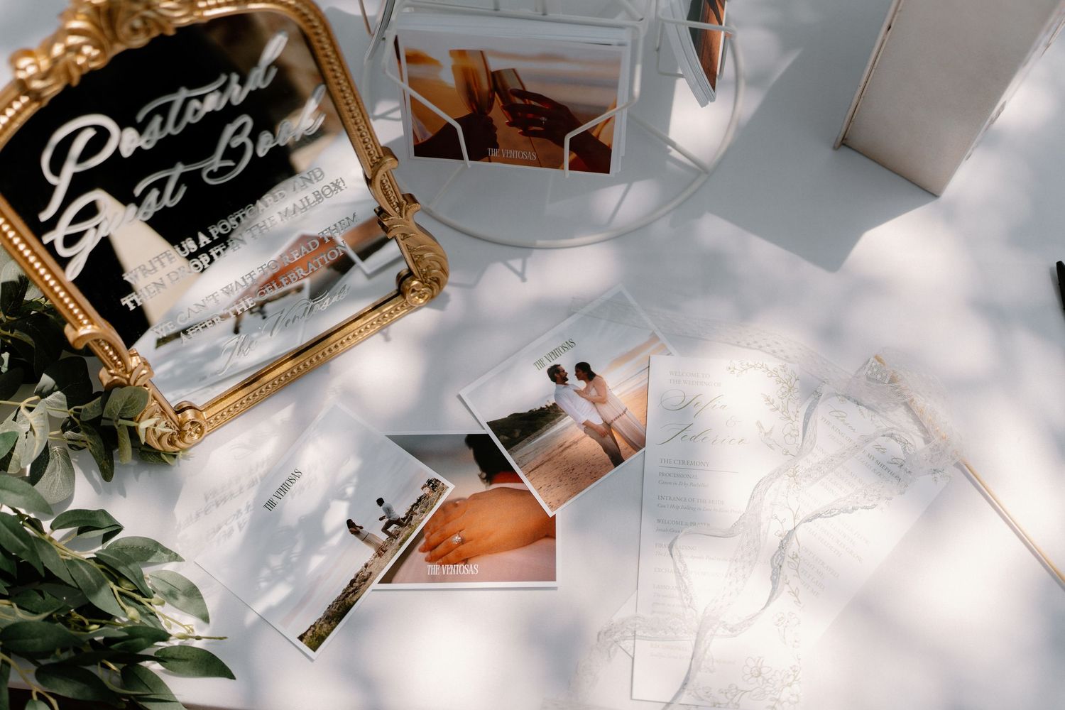 Flat lay of wedding photos, a postcard guest book mirror sign, and ribbon on a white surface.