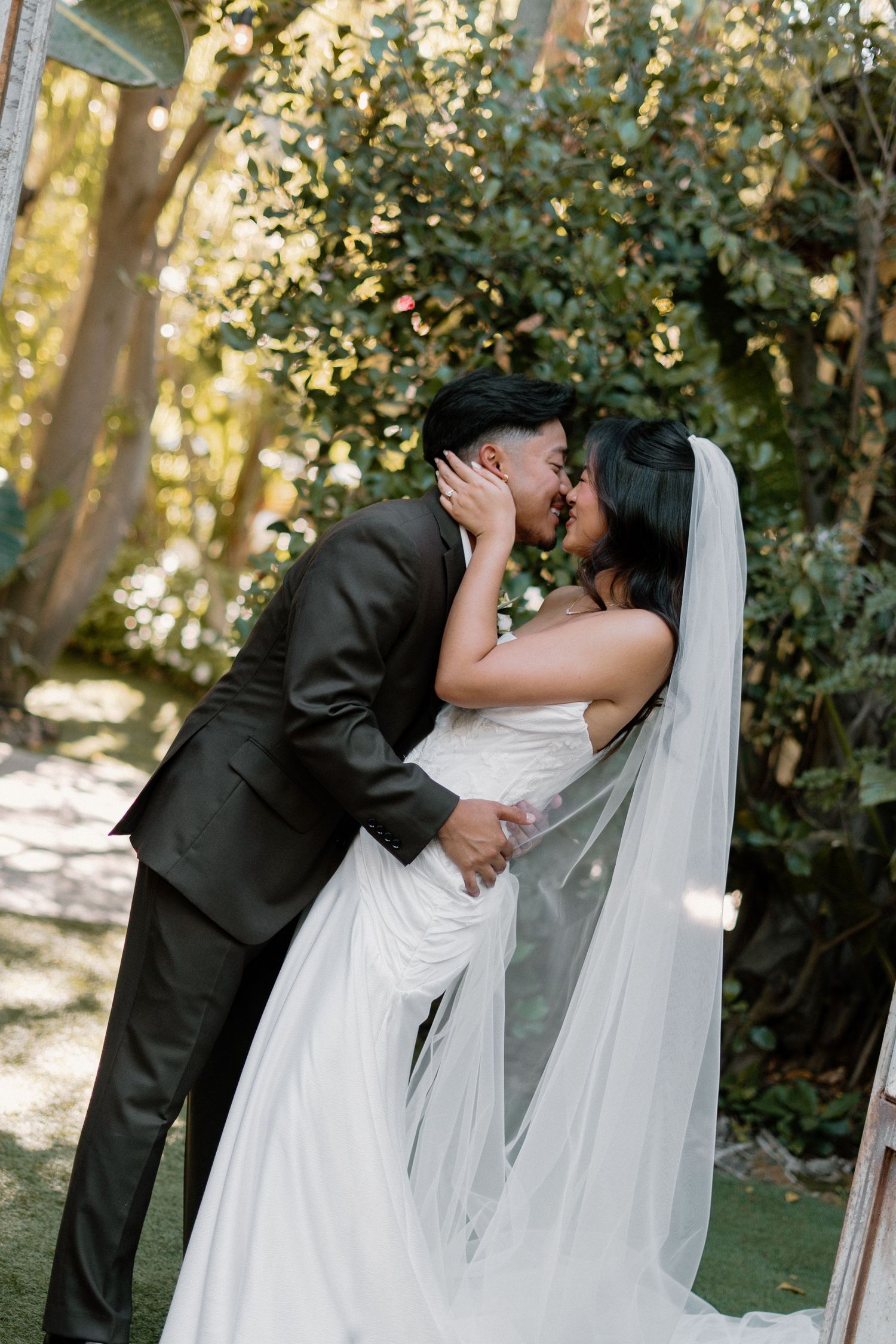 Bride and groom share first kiss at outdoor garden wedding ceremony surrounded by lush green trees.