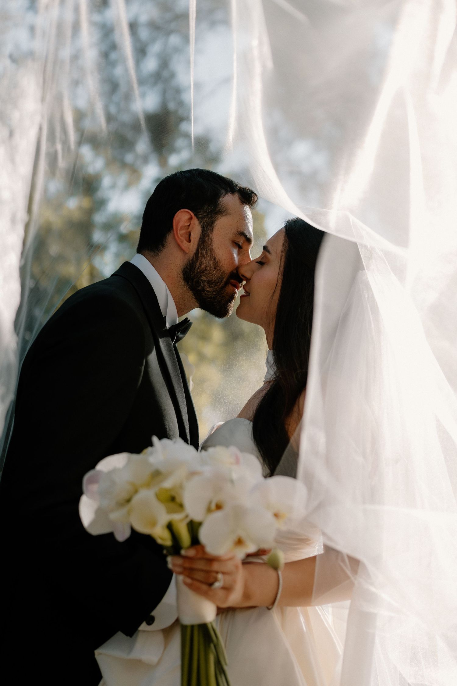 A bride and groom share a romantic kiss under a flowing white veil with a white bouquet.