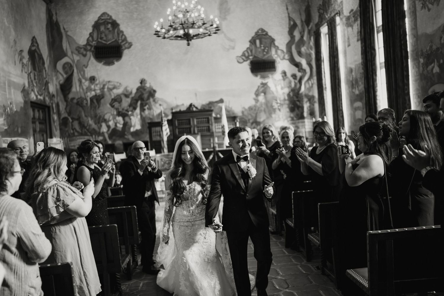 Bride and groom walk down the aisle of a grand mural-adorned church in black and white.