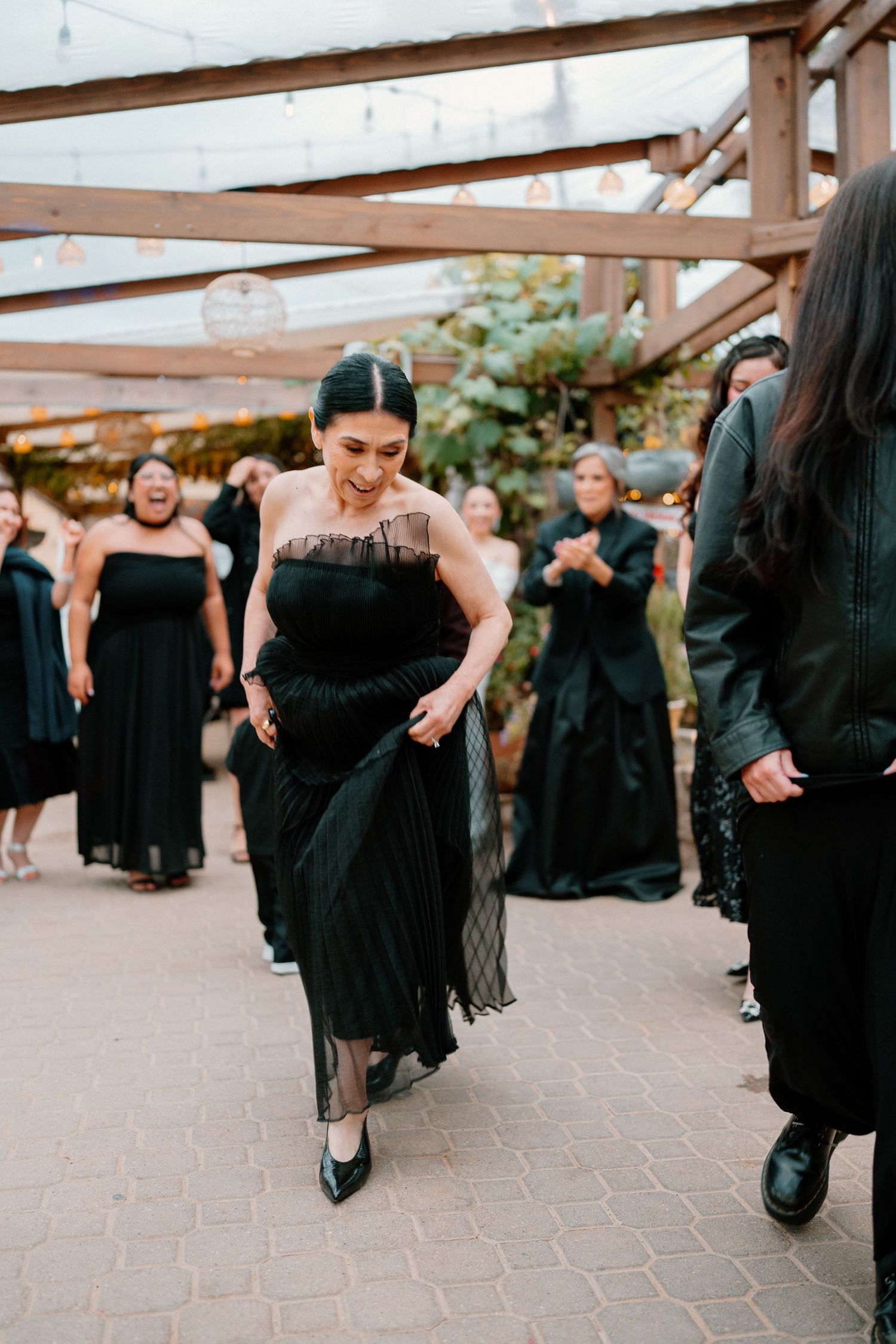 Bridesmaid in black strapless gown walking through a cheering crowd at an outdoor wedding reception.