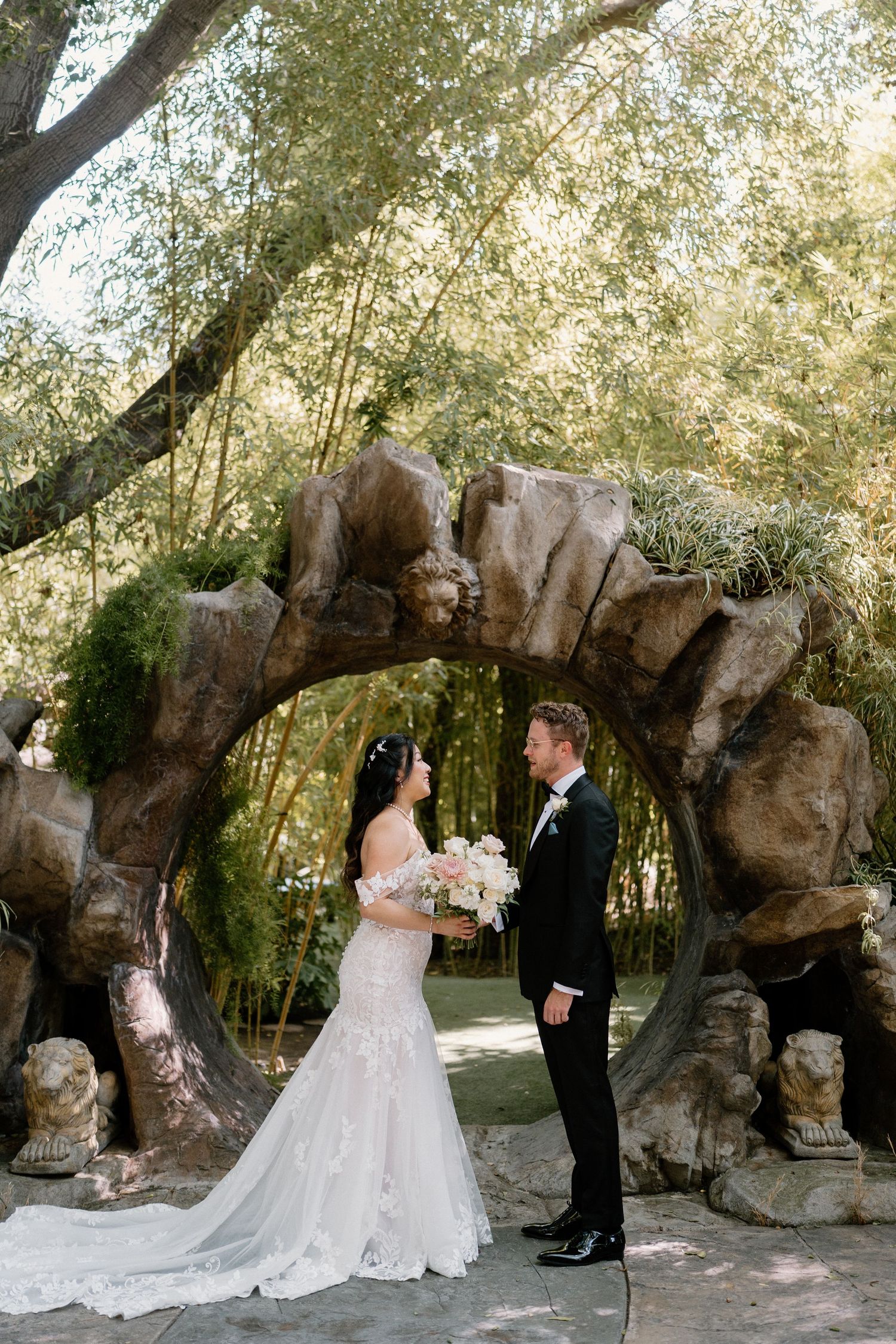 Bride and groom stand beneath a rustic stone arch surrounded by bamboo trees in a lush garden wedding.