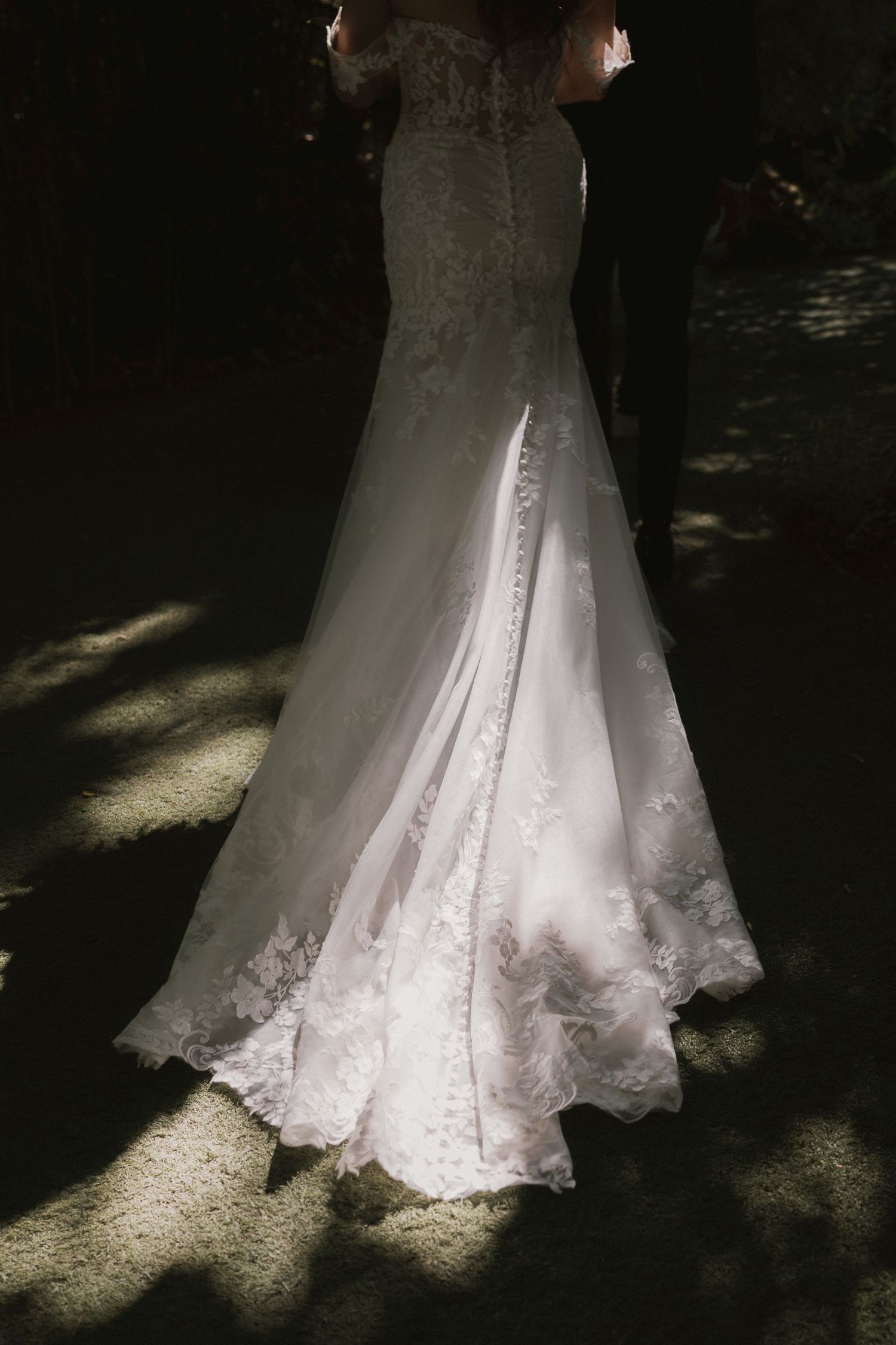 Elegant lace mermaid wedding dress with long train illuminated by dappled sunlight outdoors.