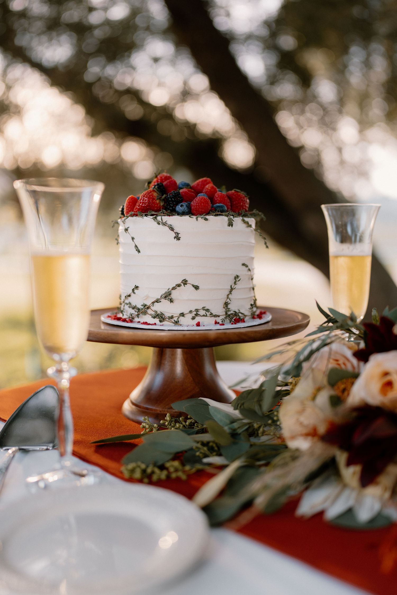 Elegant white wedding cake topped with fresh berries on wooden stand with champagne flutes outdoors.