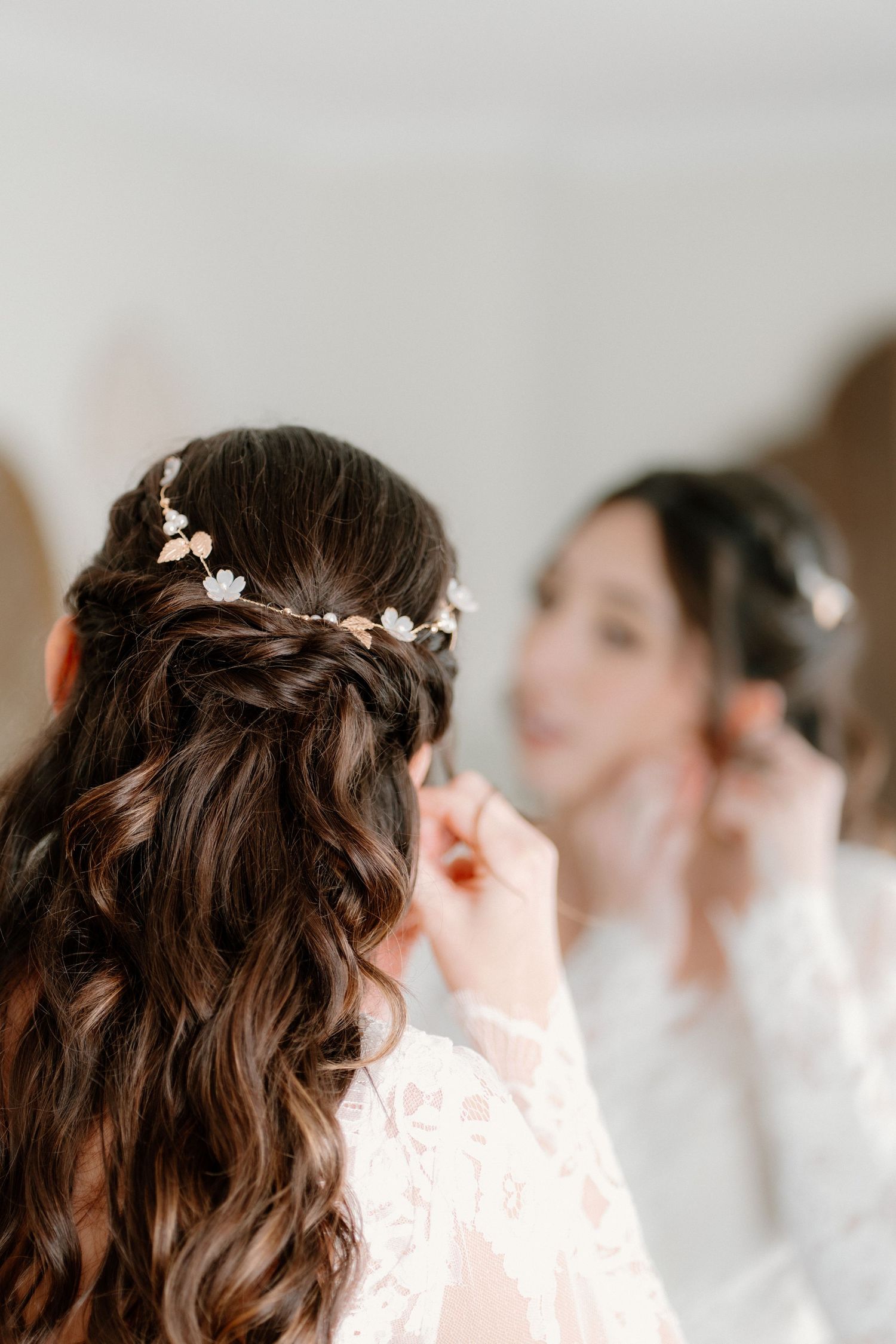 A bride with dark curly hair adorned with a delicate floral hair vine prepares in front of a mirror.