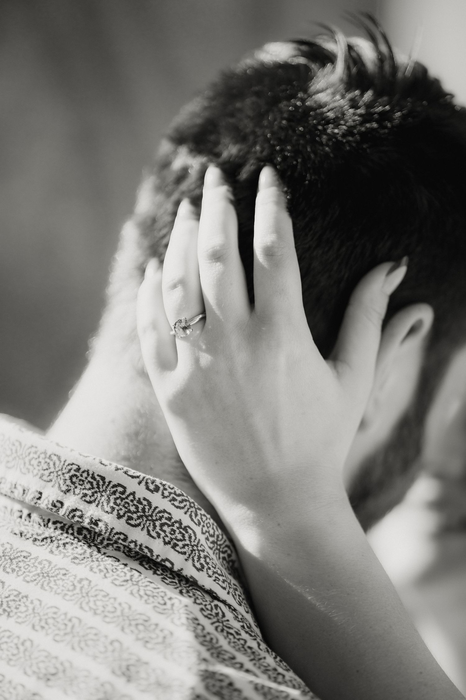 Close-up black and white photo of a womans hand with engagement ring resting on a mans head.