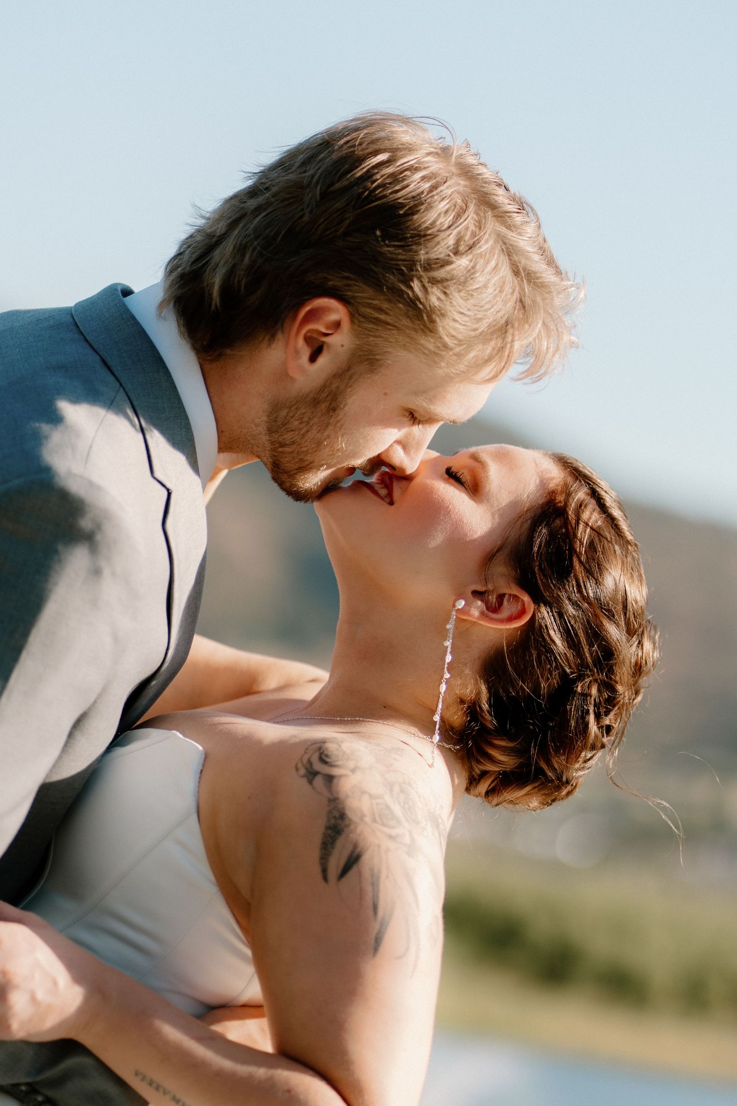 Couple shares romantic kiss outdoors on sunny wedding day, bride with floral tattoo in white dress.