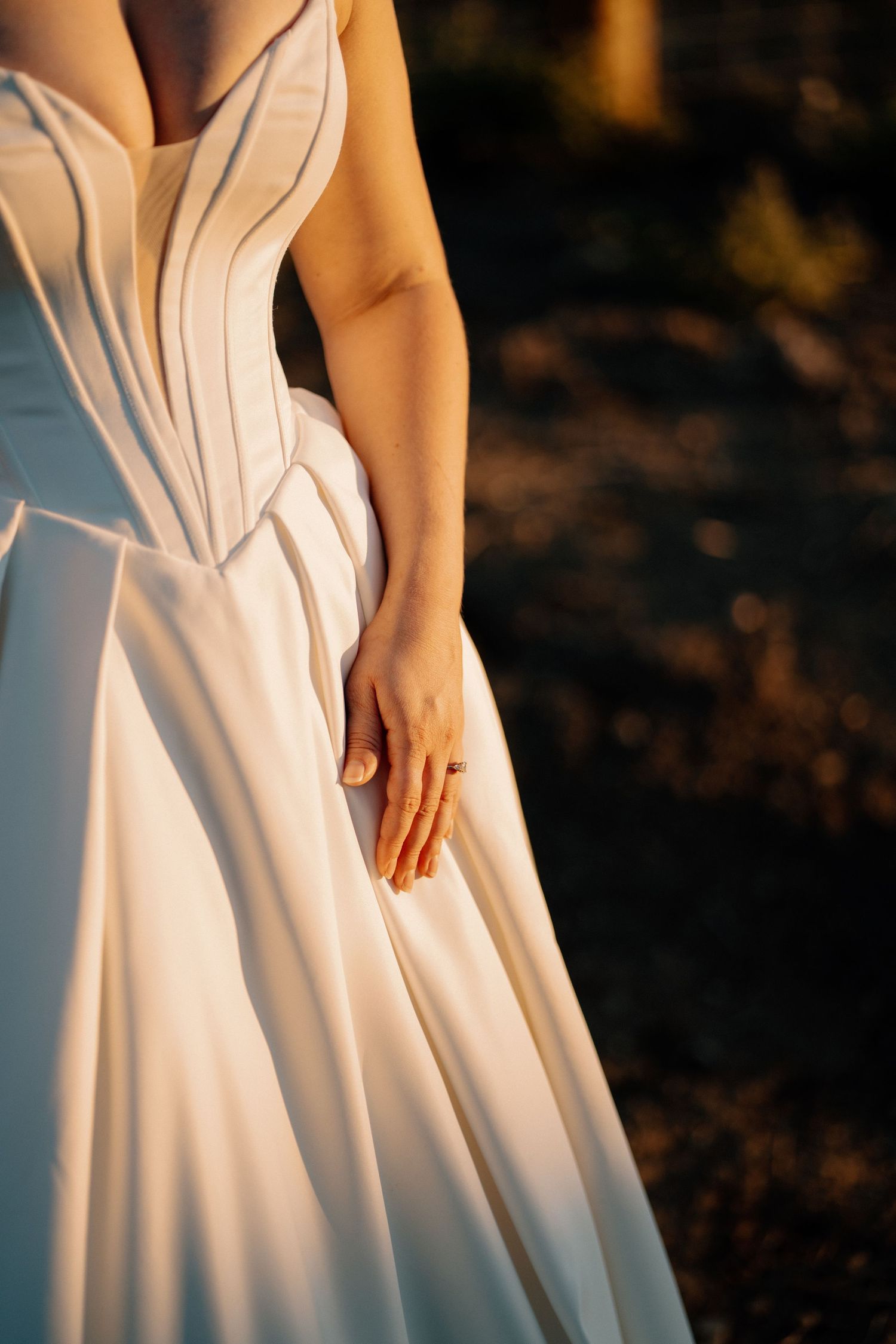 Close-up of a bride in a structured white corset gown with her hand resting on the skirt at sunset.