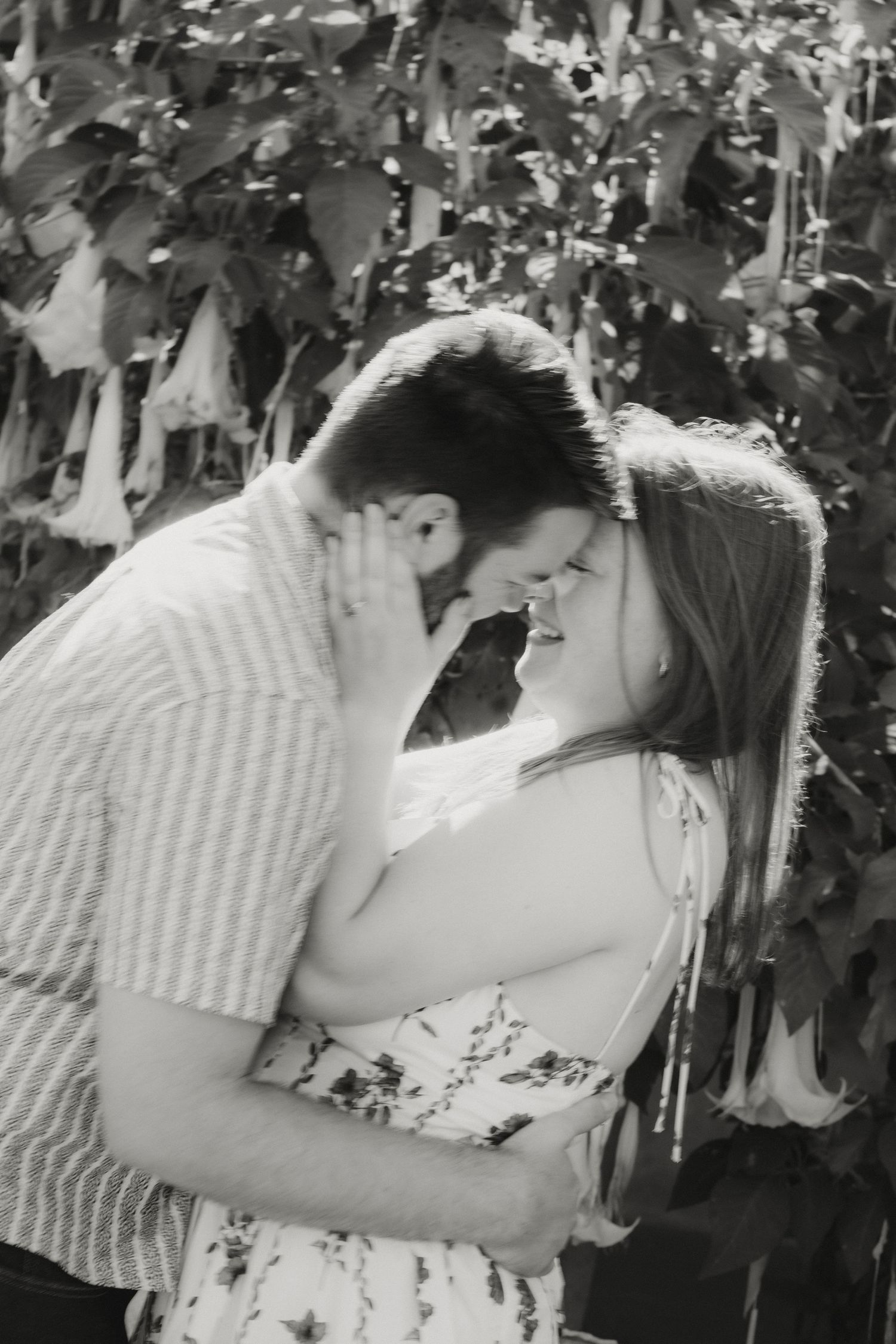 Black and white romantic photo of couple nearly kissing, embracing among lush foliage in soft light.