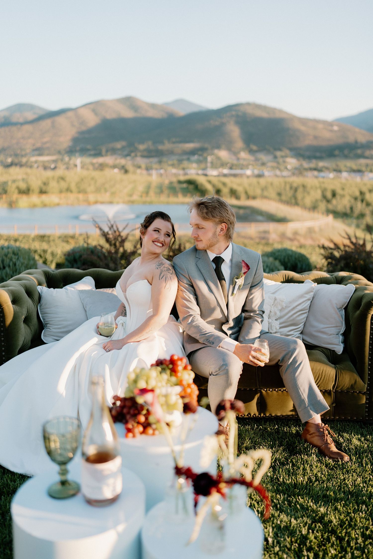 Bride and groom sit together on a green tufted sofa outdoors with stunning mountain and lake views behind them.