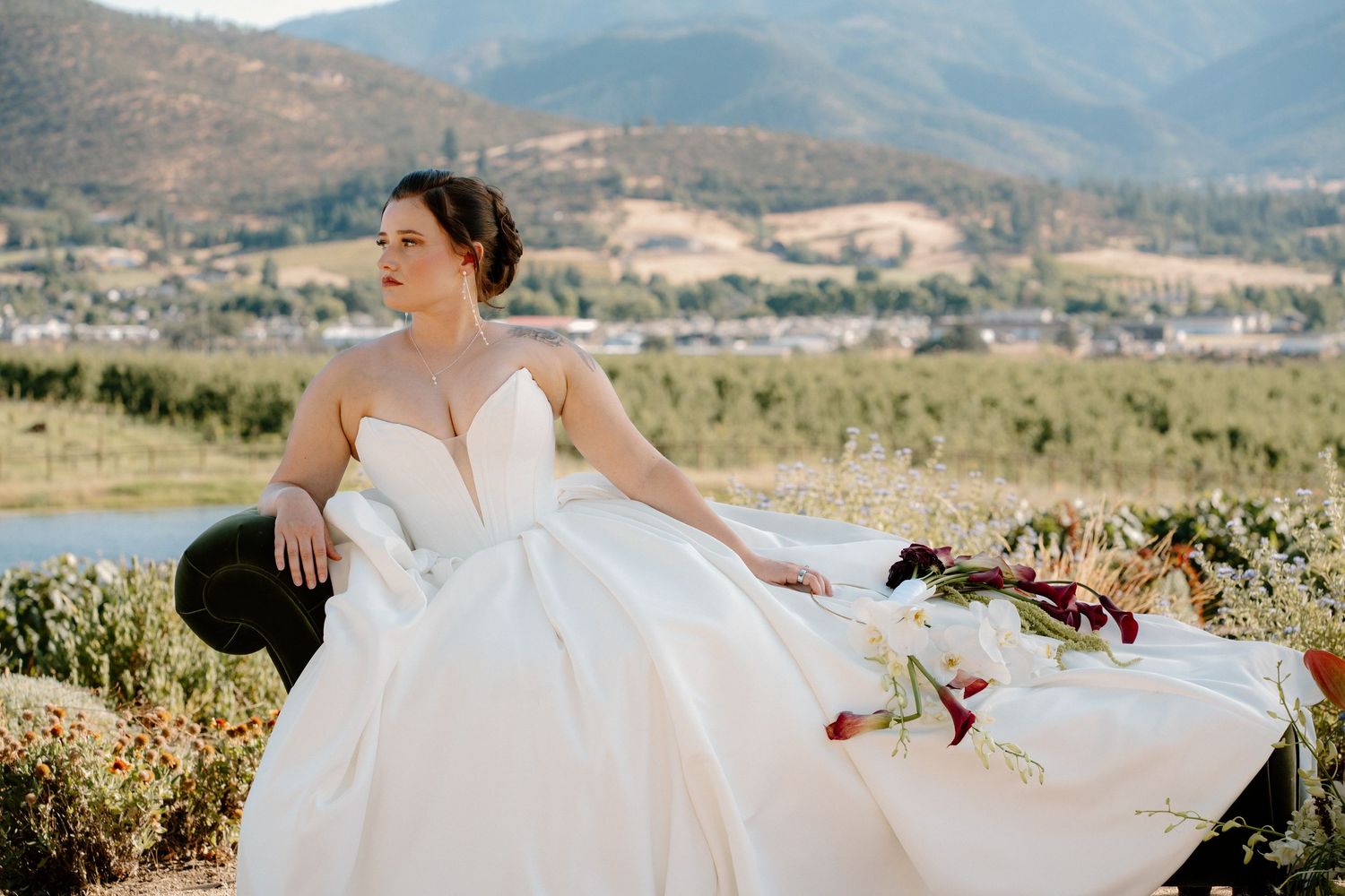 Bride in a white ball gown sits gracefully outdoors with mountain and vineyard views in the background.