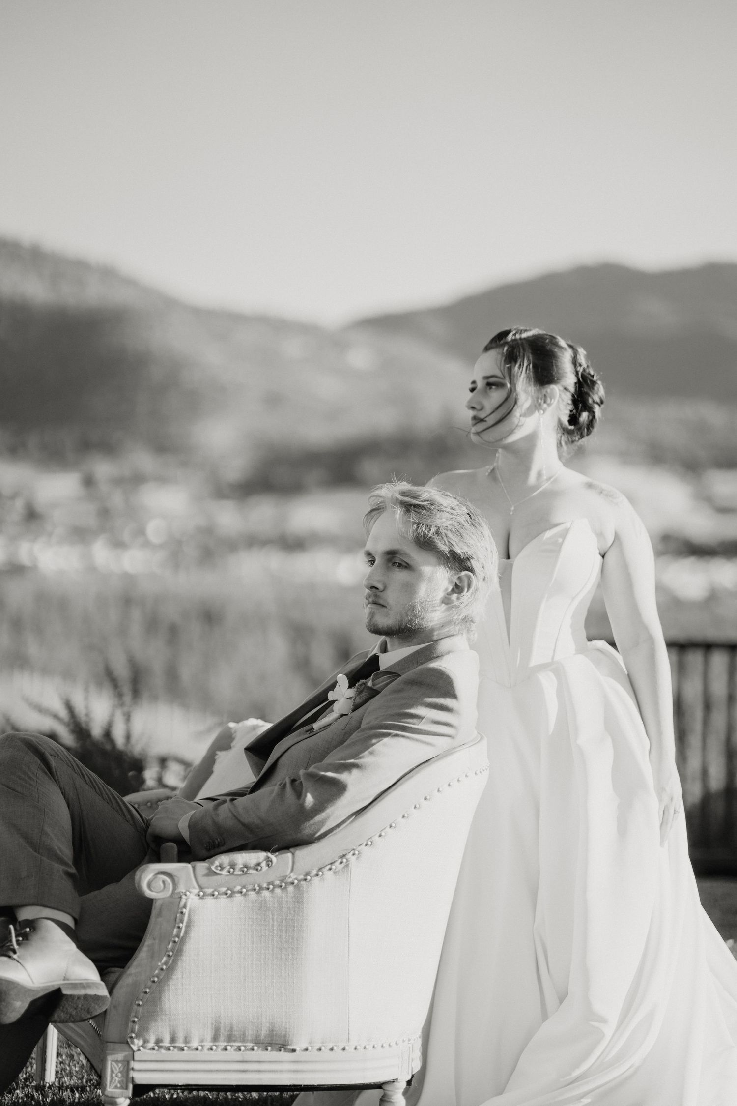 Elegant black and white portrait of bride and groom posing with vintage chair and mountain scenery.