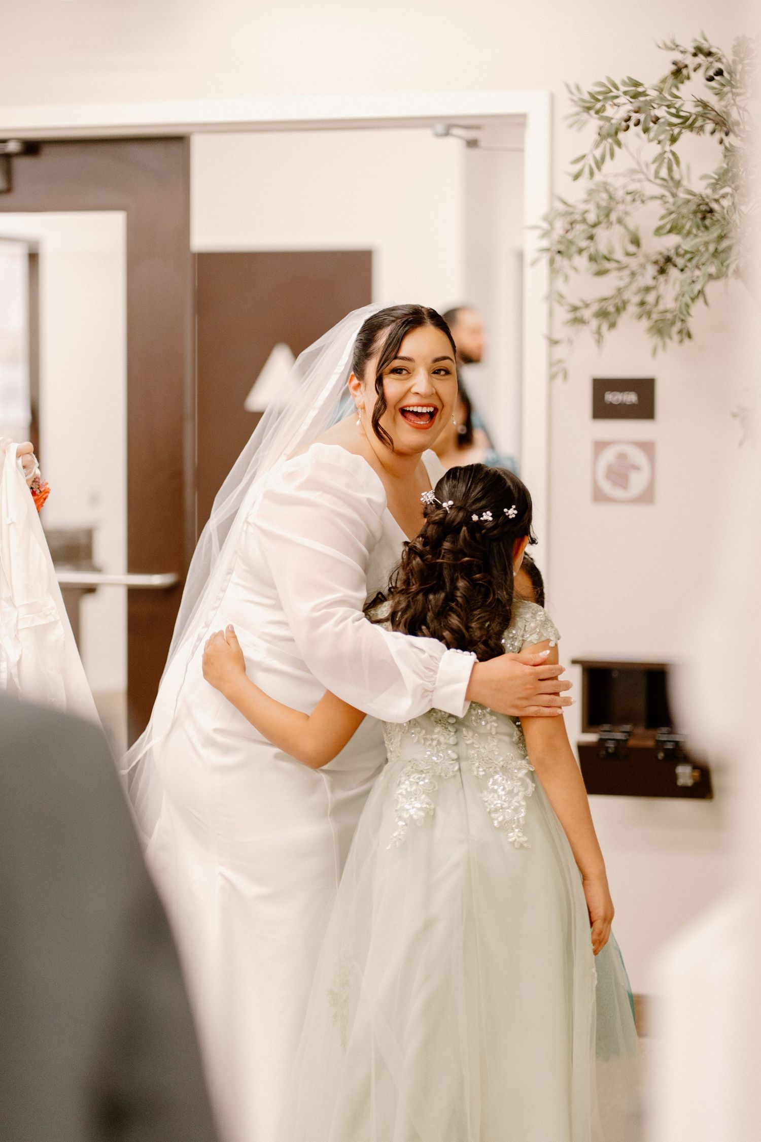 Bride in white gown and veil hugging a young flower girl in a sage green dress at a wedding.