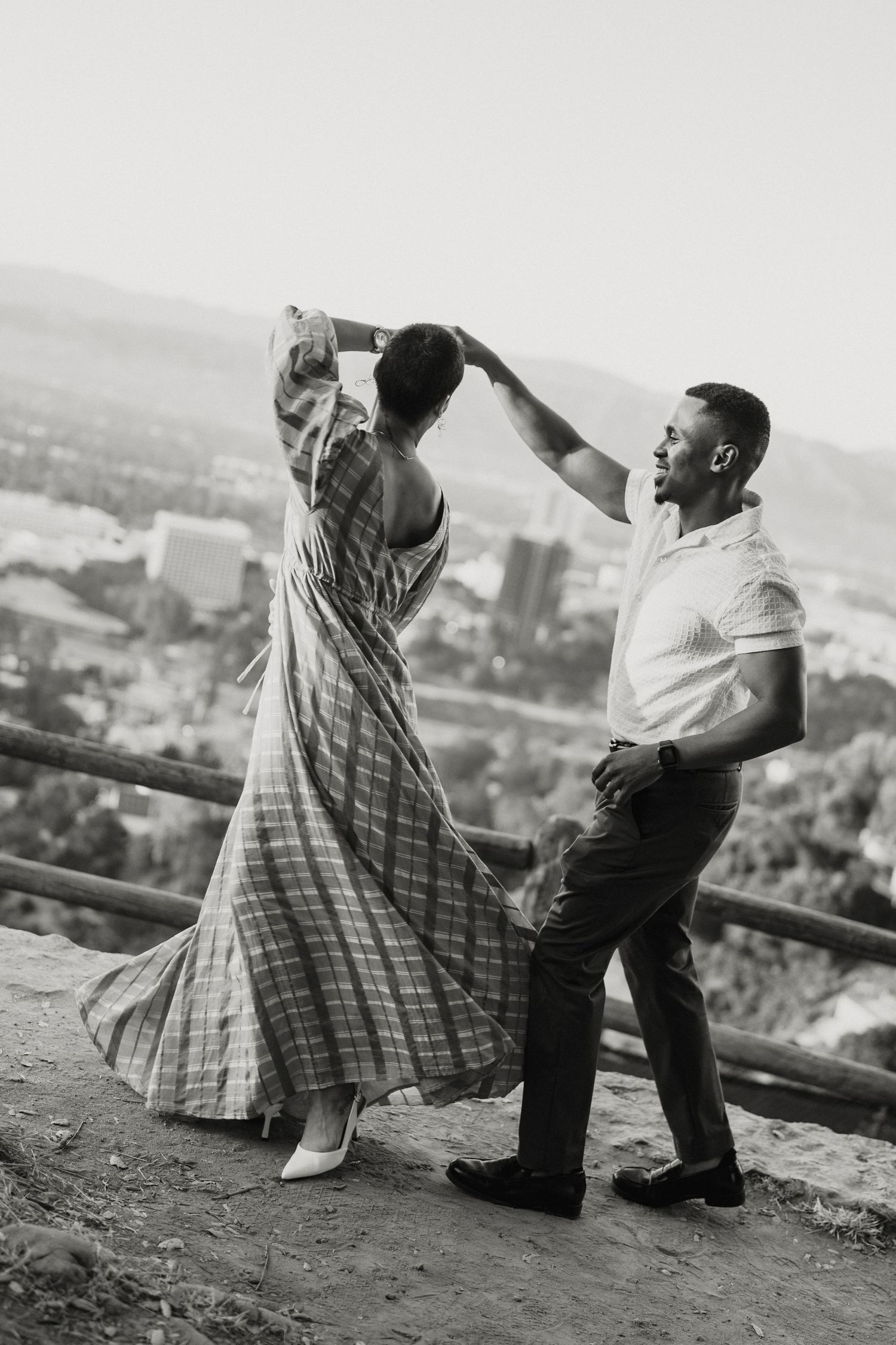 Black and white photo of a couple dancing outdoors with a city skyline backdrop, man spinning woman in plaid dress.