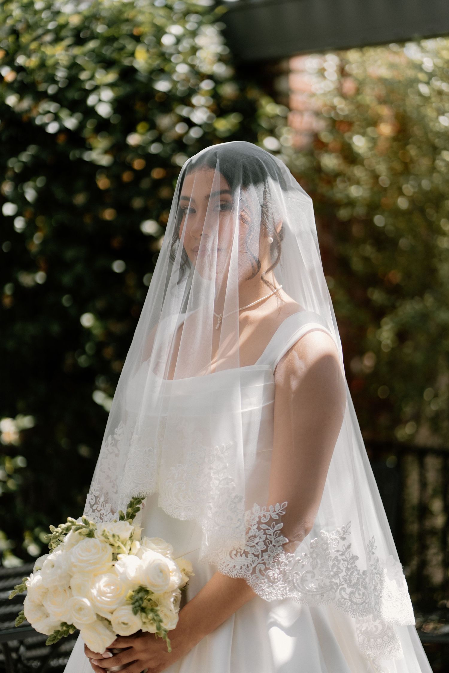 Beautiful bride wearing lace-trimmed veil over face holding white peony bouquet in garden setting.