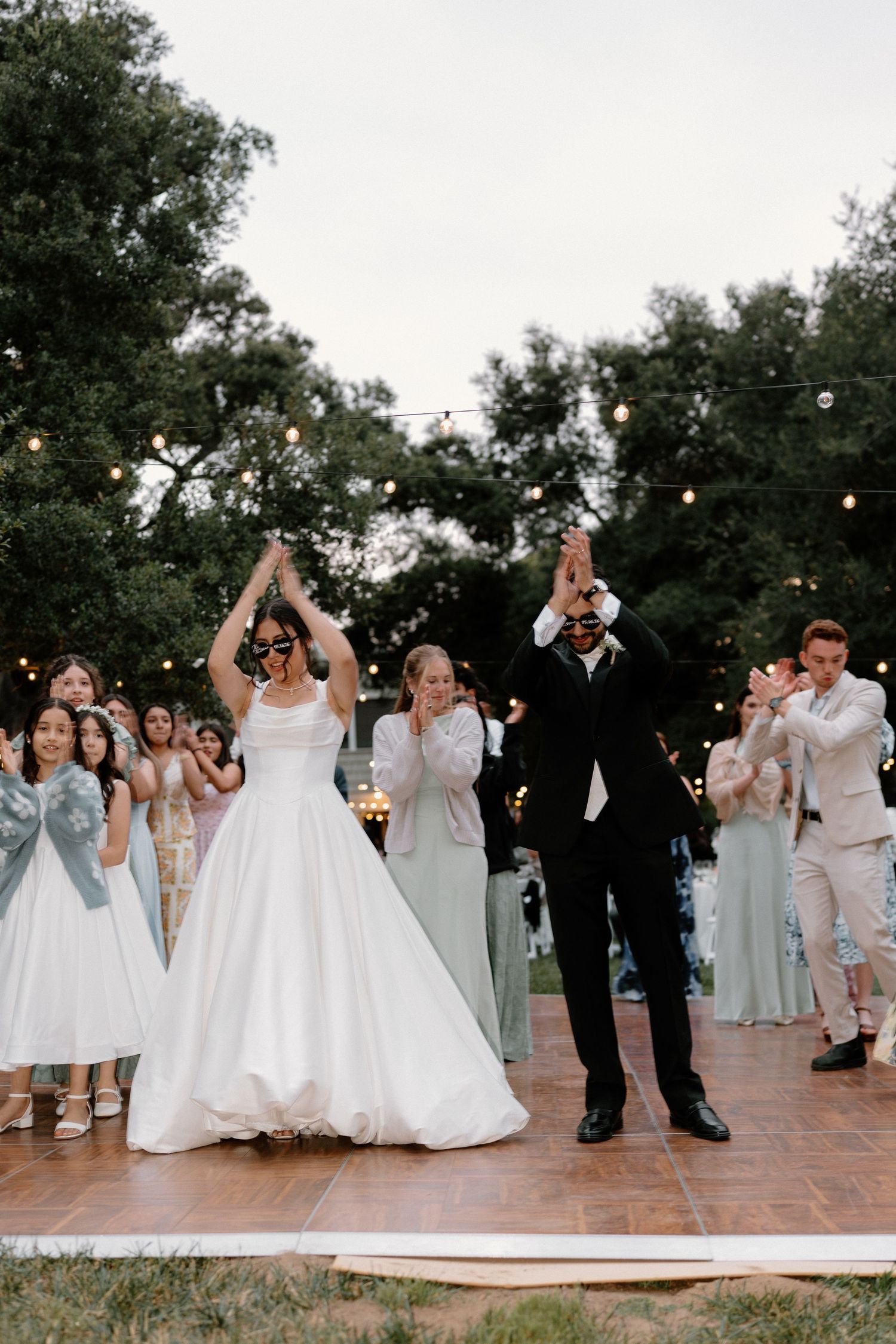 Bride and groom raise hands celebrating grand entrance at outdoor wedding reception with string lights.
