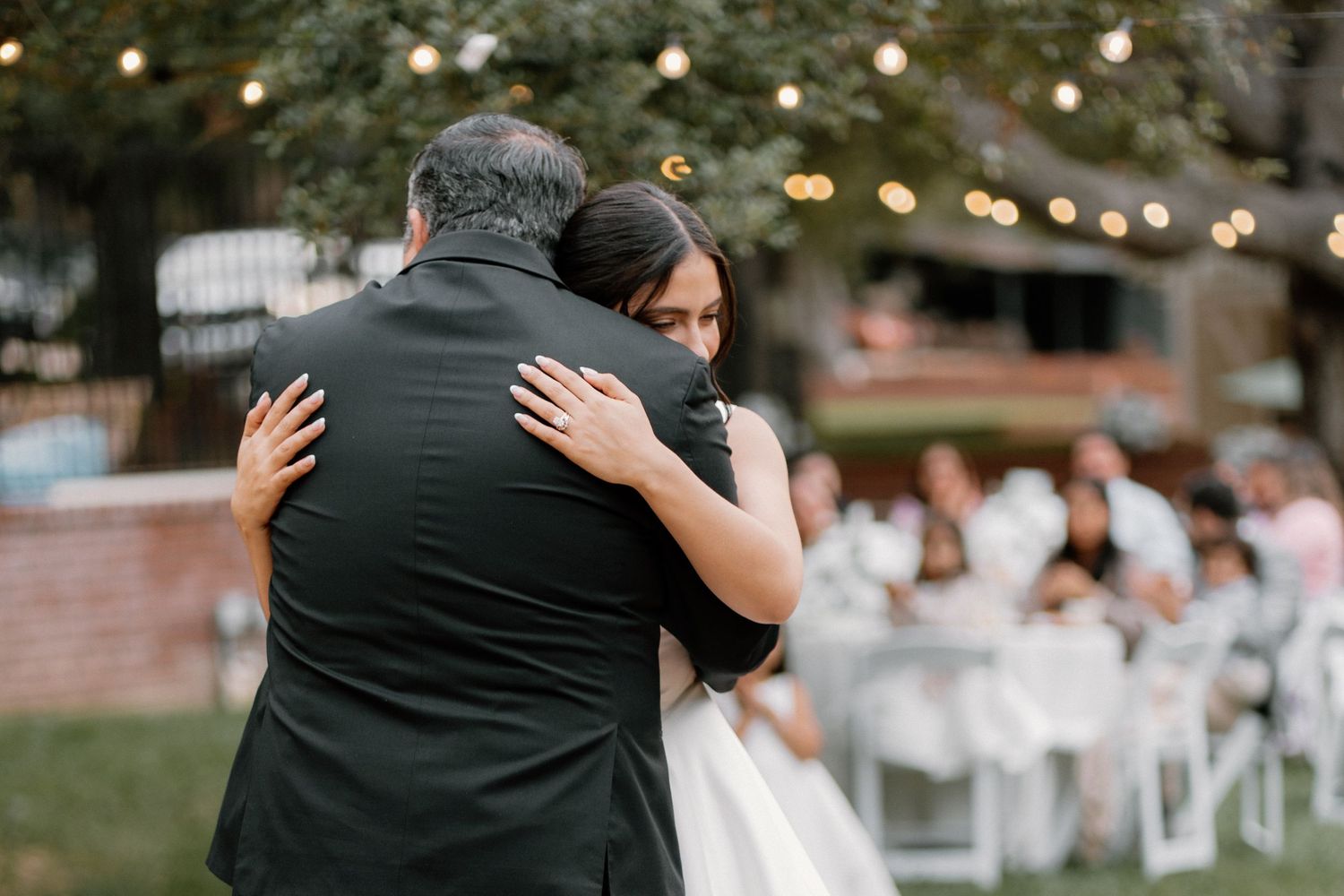 Bride and father share emotional first dance embrace at outdoor wedding reception with string lights.