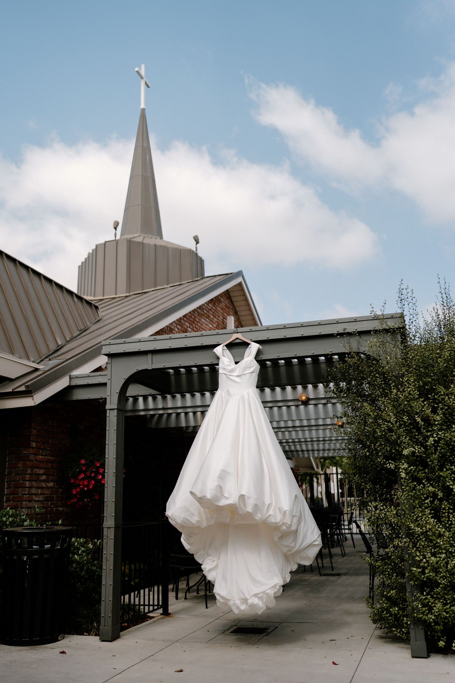 White wedding dress hanging under a pergola with a church steeple and cross visible against a blue sky.