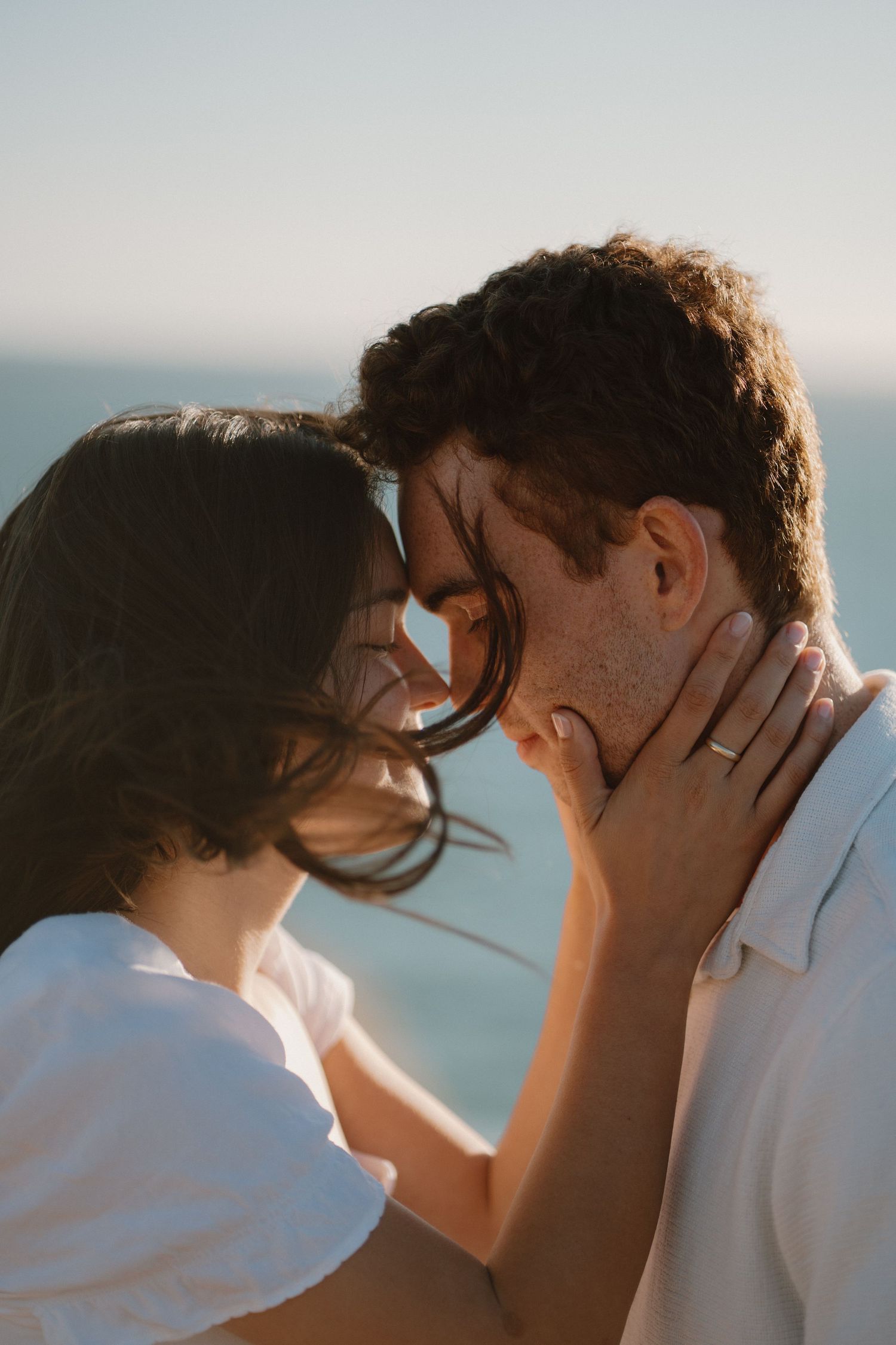 Romantic couple touching foreheads with woman's hand on man's neck showing engagement ring by ocean.