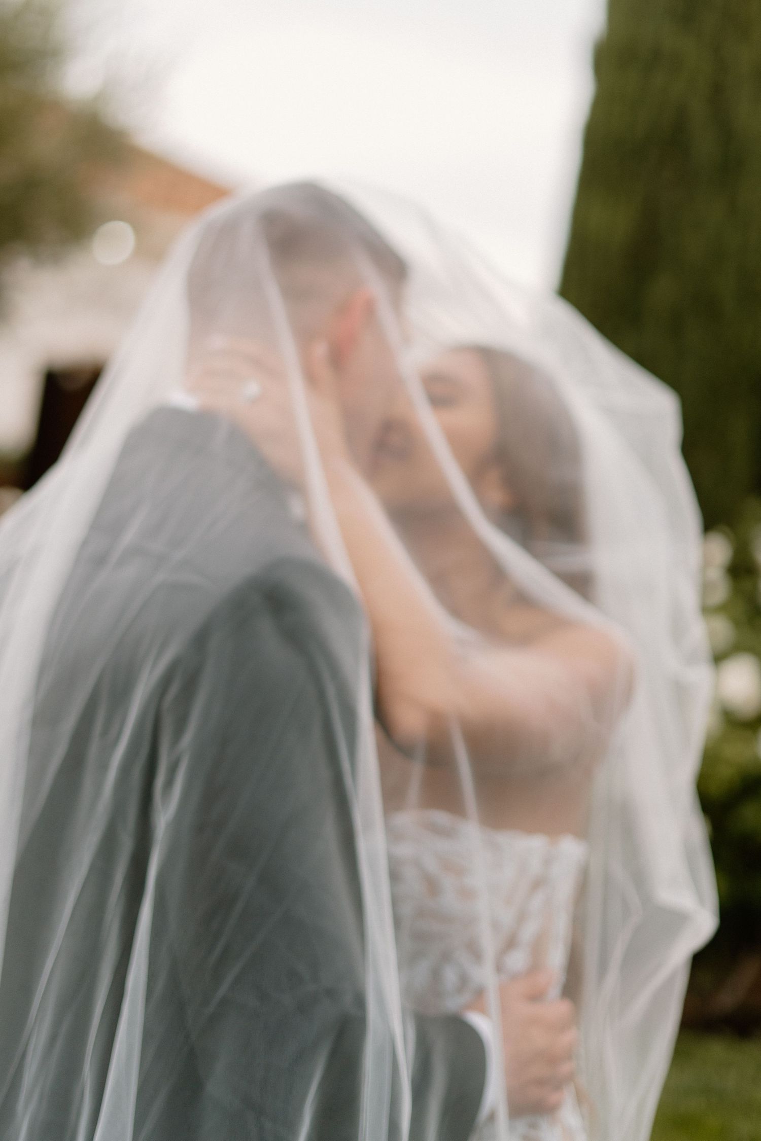 Romantic blurred image of bride and groom sharing a kiss under a flowing white wedding veil outdoors.