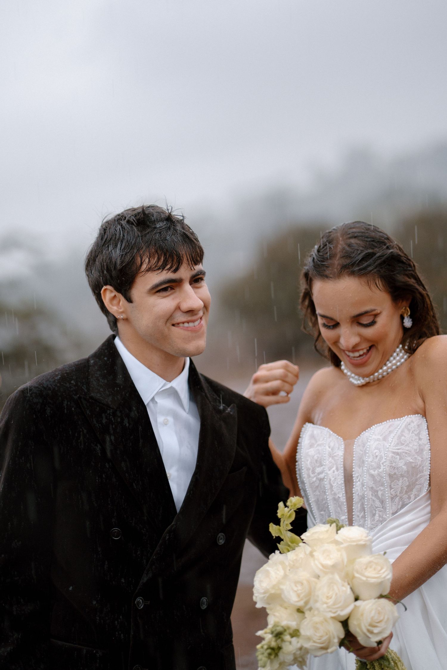 Smiling groom in black coat stands beside laughing bride holding white rose bouquet in the rain on their wedding day.