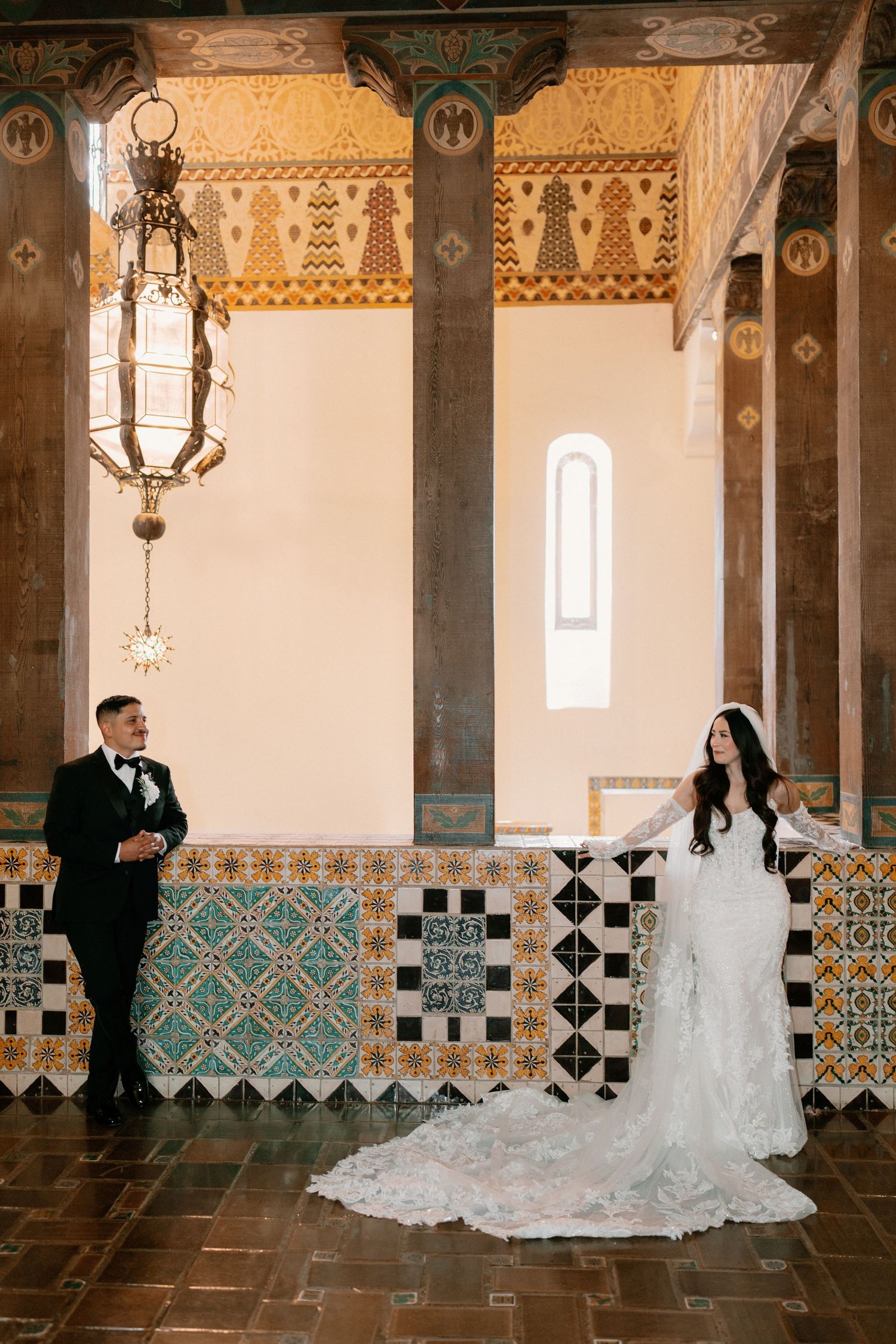 Bride and groom posing in ornate hall with colorful mosaic tiles and hanging lantern.