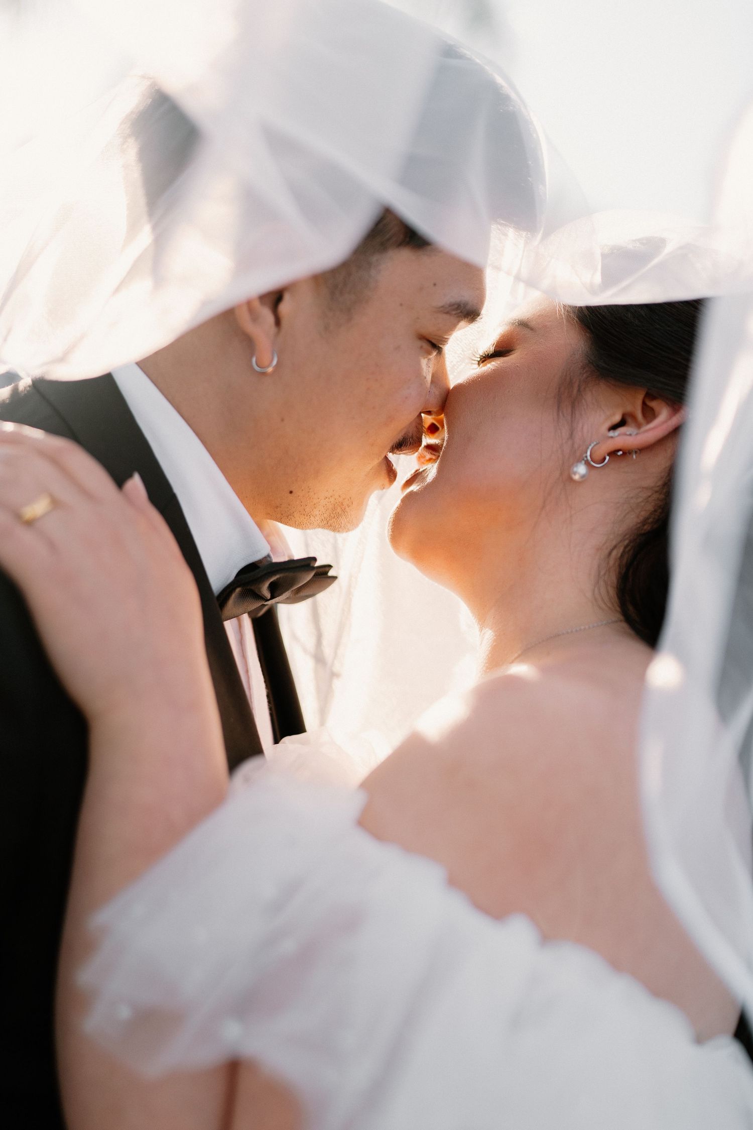 Bride and groom sharing a romantic kiss under a flowing white veil with soft backlight on their wedding day.