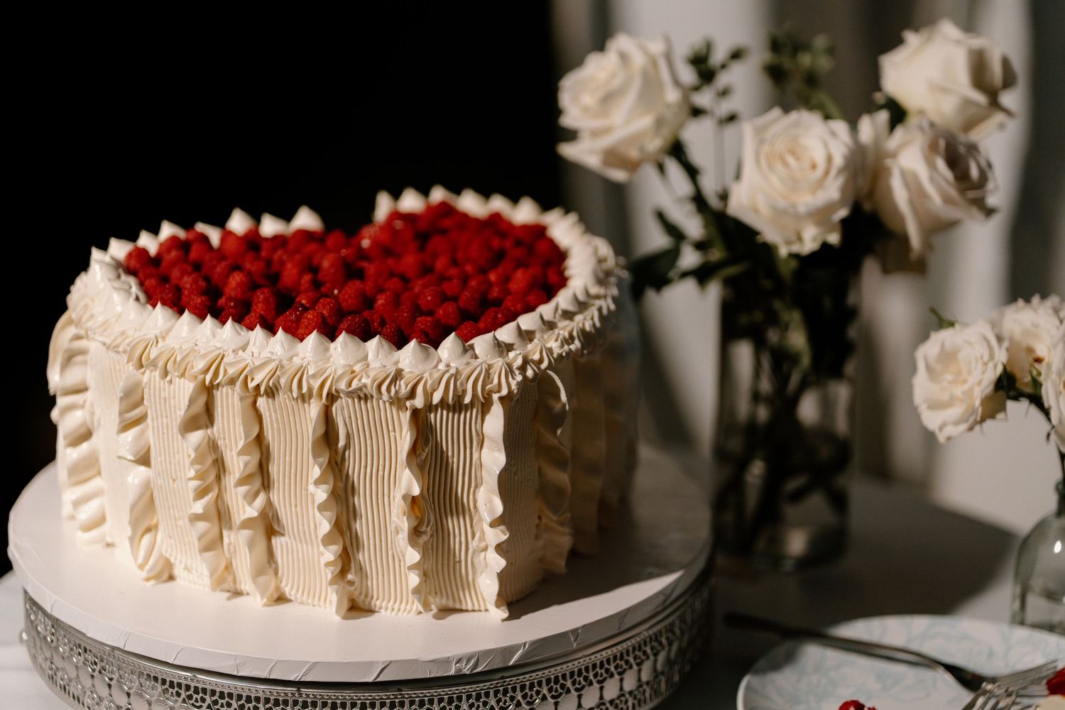 Elegant white wedding cake topped with fresh raspberries displayed beside white roses.