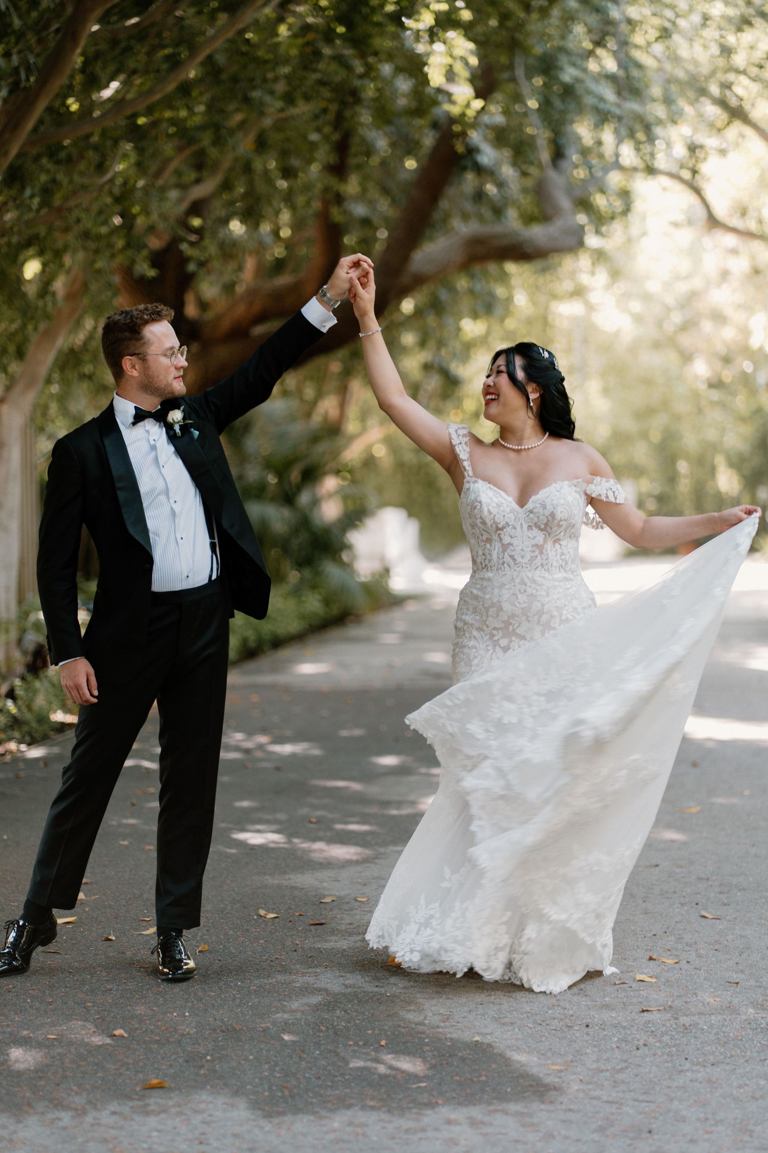 Bride in lace off-shoulder gown twirls with groom in black tuxedo on a tree-lined path.
