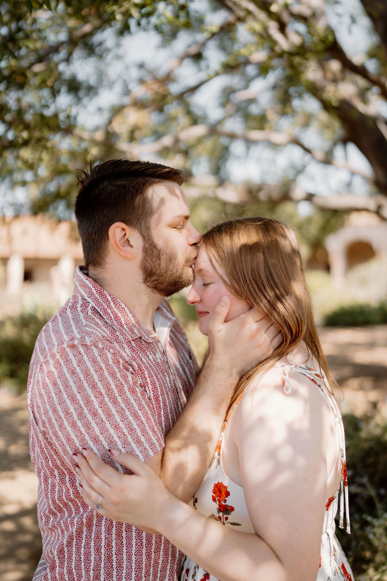 Couple sharing a kiss under a tree in dappled sunlight during outdoor engagement session.