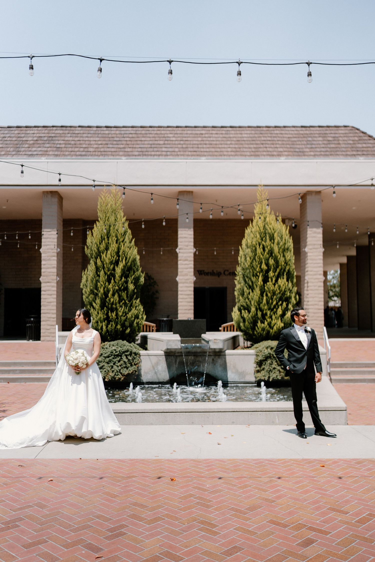 Bride and groom stand apart near a fountain courtyard with tall cypress trees and string lights at their wedding venue.
