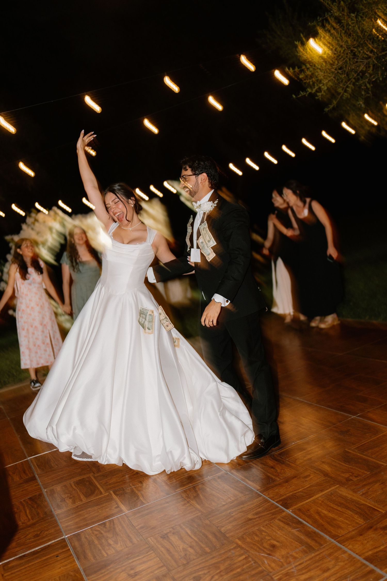 Bride in white gown raises arm joyfully while dancing with groom under string lights at night reception.