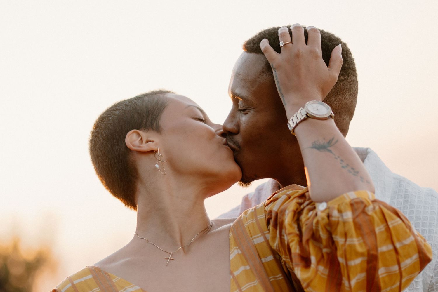 A couple shares a romantic kiss at sunset, with the woman in a yellow floral top and the man wearing a watch.