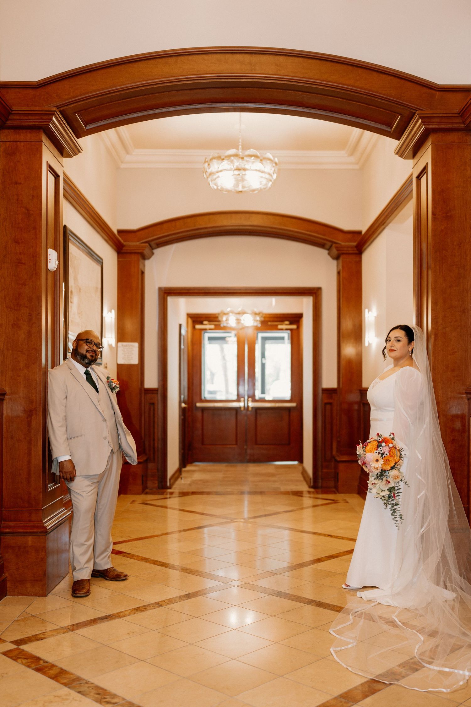 Bride and groom pose apart in ornate wood-paneled hall, bride holding vibrant orange bouquet.