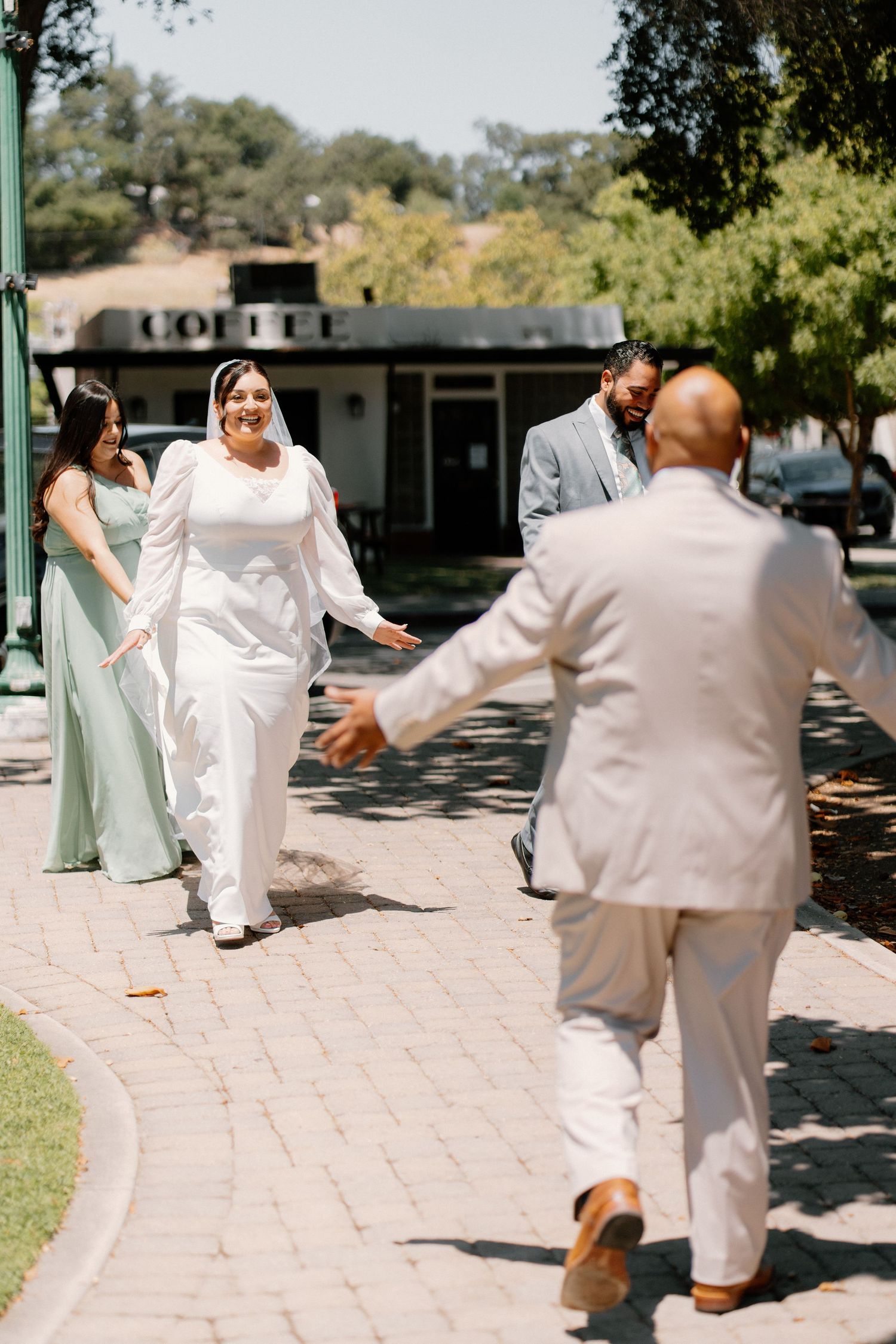 Bride in white gown walks toward groom on brick path, with bridesmaids in sage green dresses at outdoor wedding.